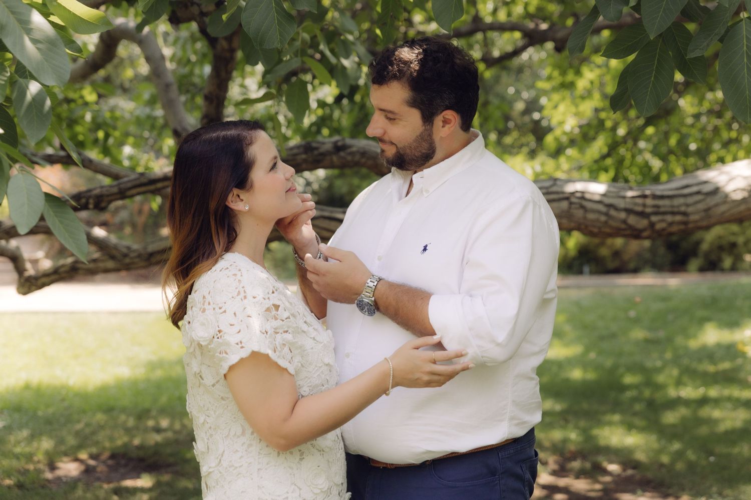 Man tenderly touches womans face as couple shares an intimate moment under leafy tree branches.