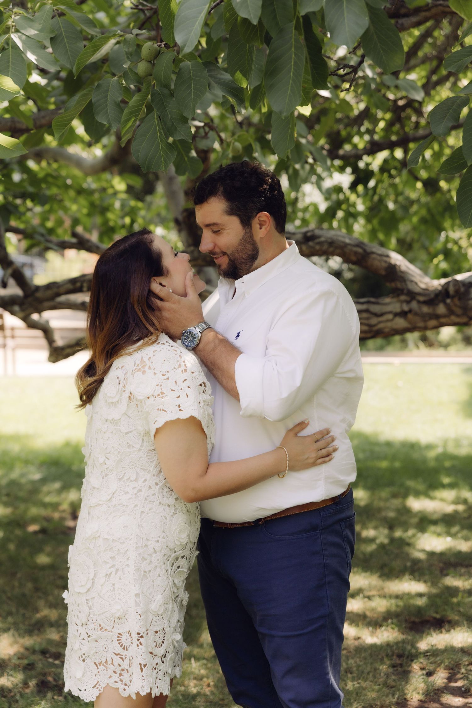 Bearded man gently holds womans face as couple stands close together in a lush green park.