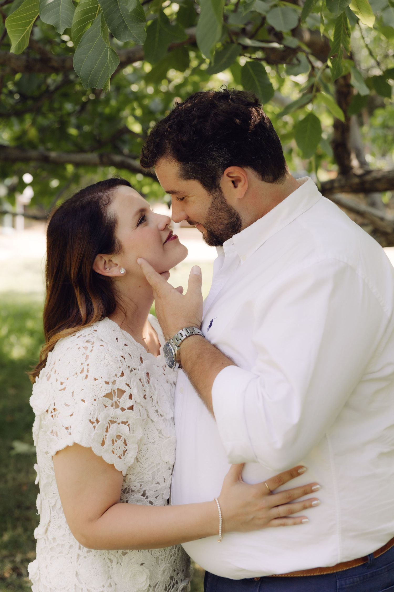 Smiling couple nearly kissing while embracing outdoors under green foliage in natural sunlight.