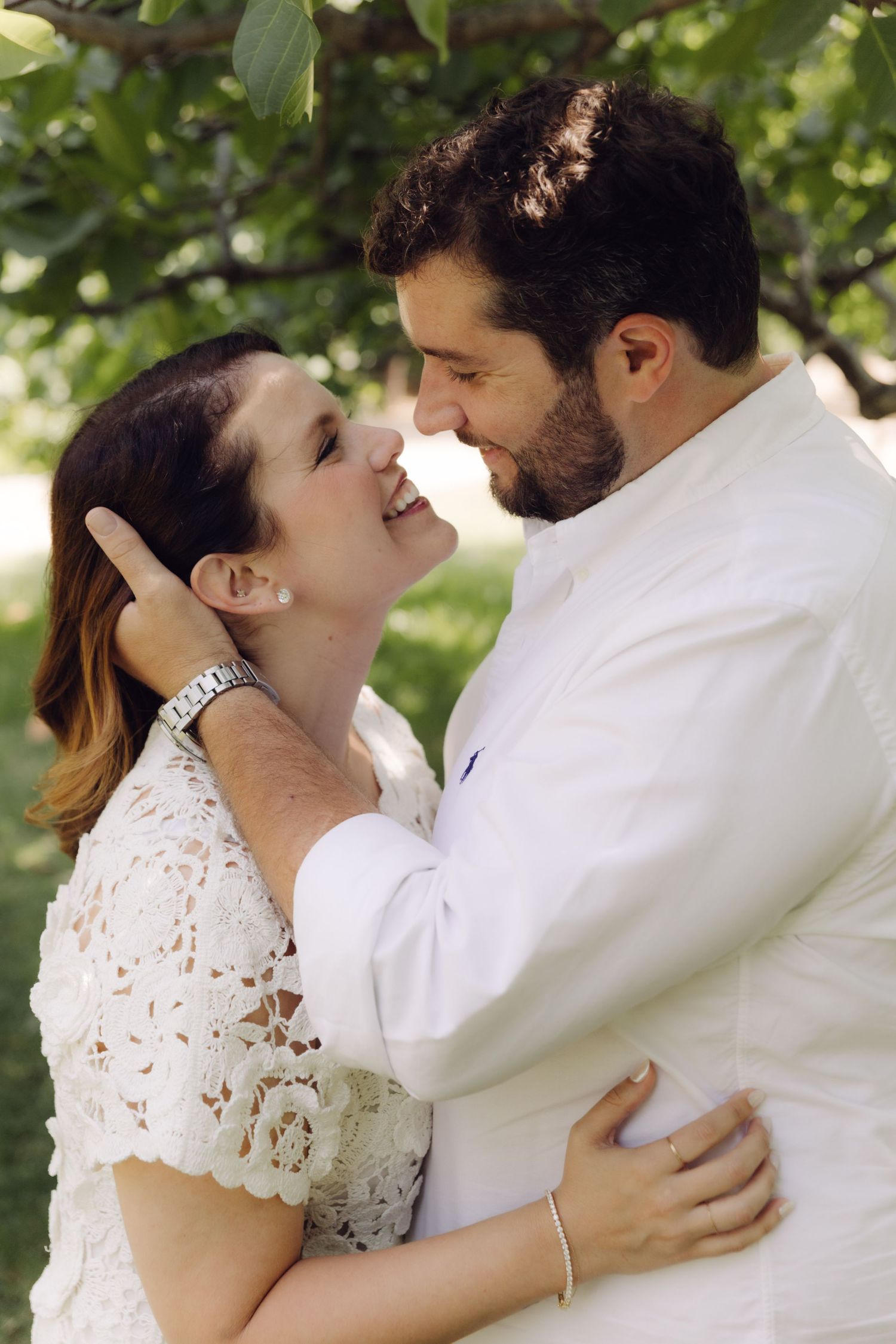 Laughing couple gazing at each other lovingly while embracing under leafy green tree branches.