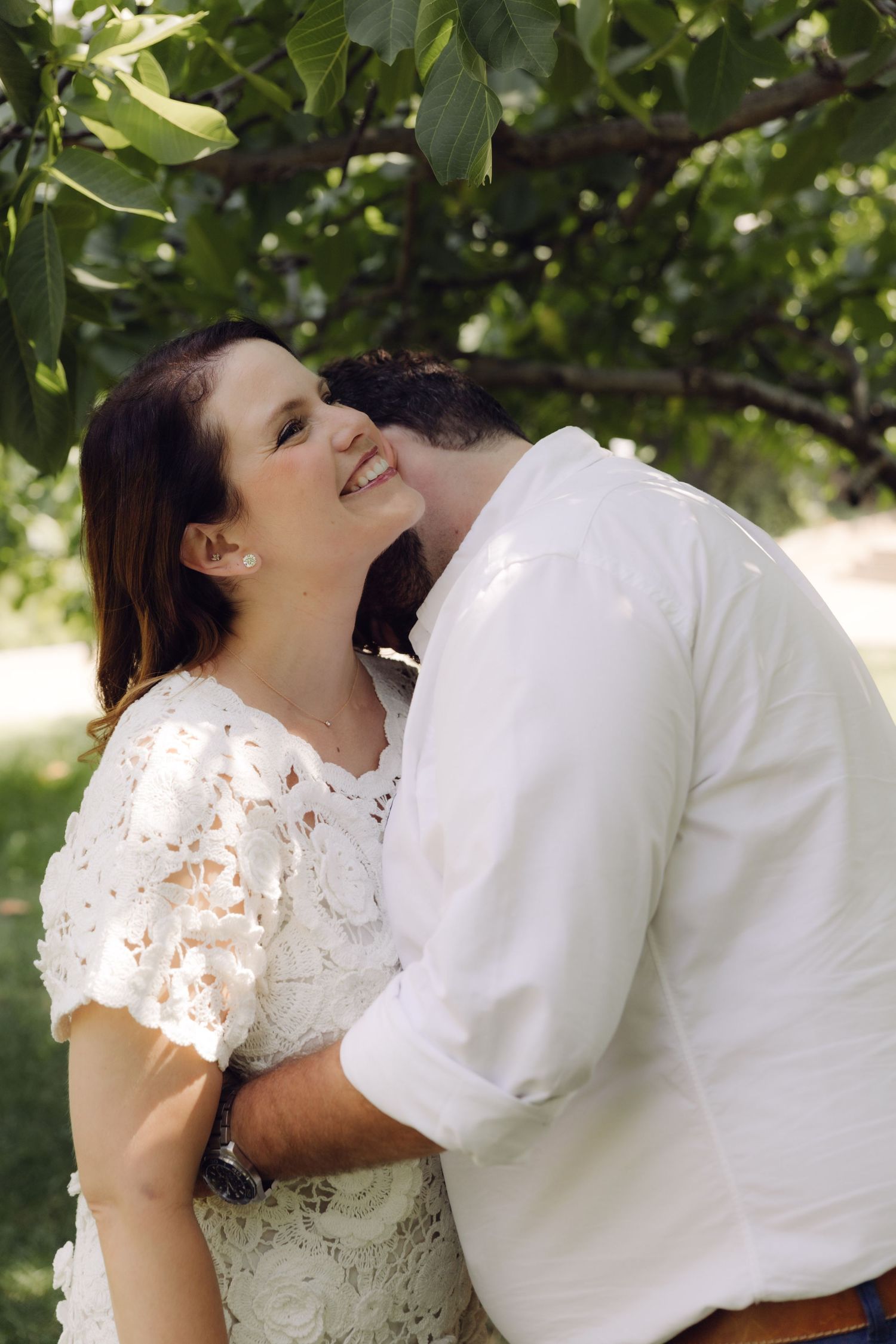 Woman smiles joyfully as man nuzzles her cheek while embracing in a sunlit green garden setting.
