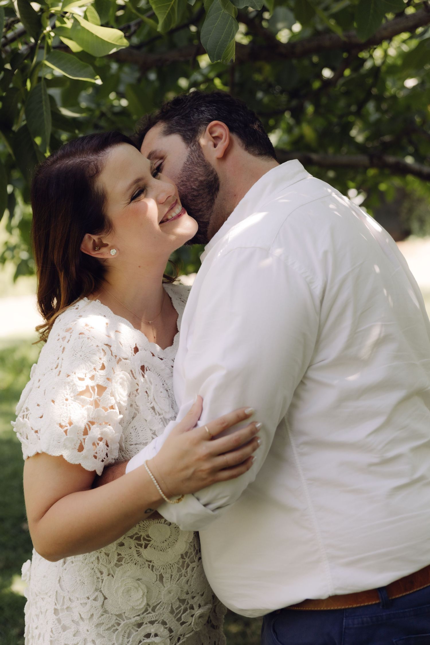 Couple sharing a gentle cheek kiss while embracing outdoors under lush green tree branches.