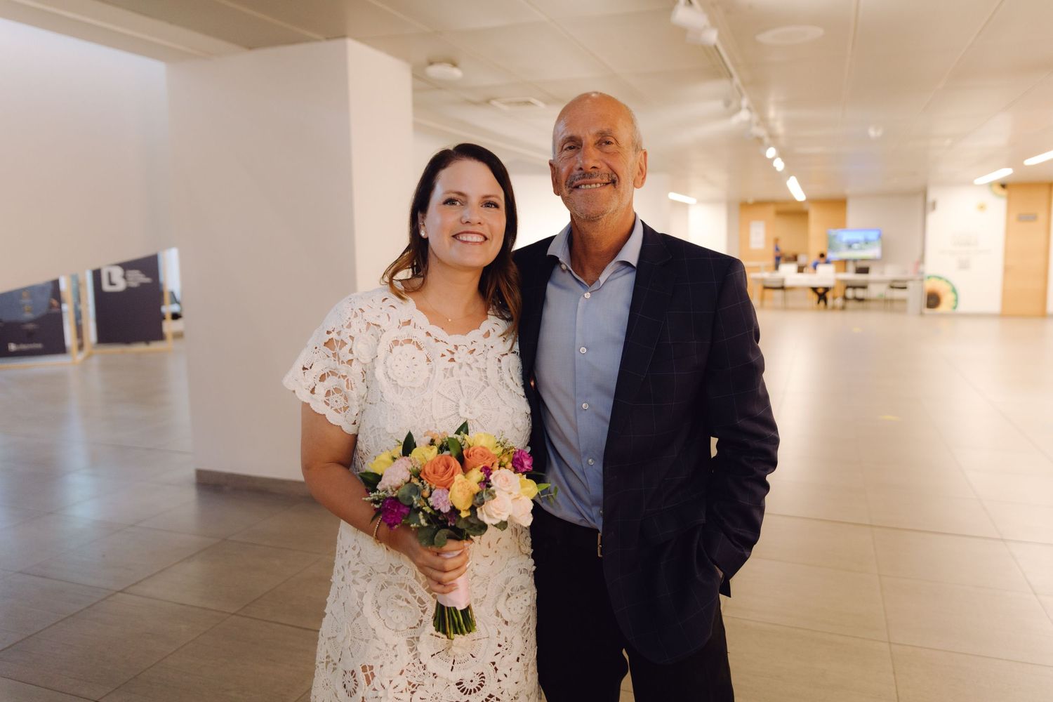 Smiling bride in white lace dress holding colorful bouquet poses with older man in navy blazer at indoor venue.
