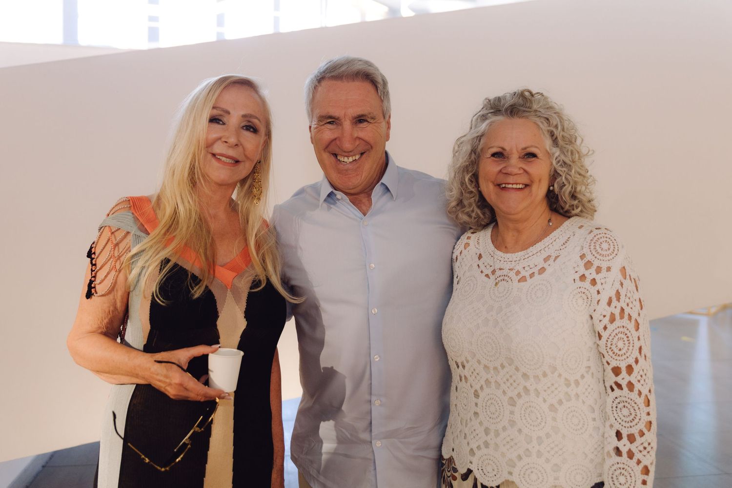 Three adults smiling together at bright indoor event, woman in black dress, man in blue shirt, woman in white lace top.