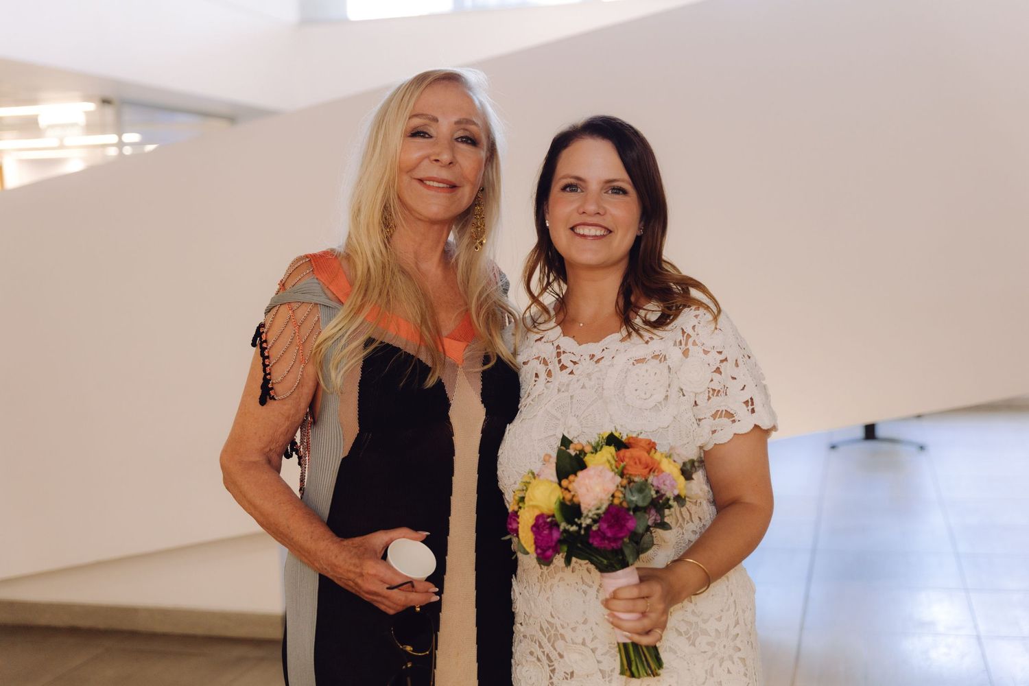 Two women smiling together at indoor event, one in black dress holding white object, other in white lace dress with bouquet.