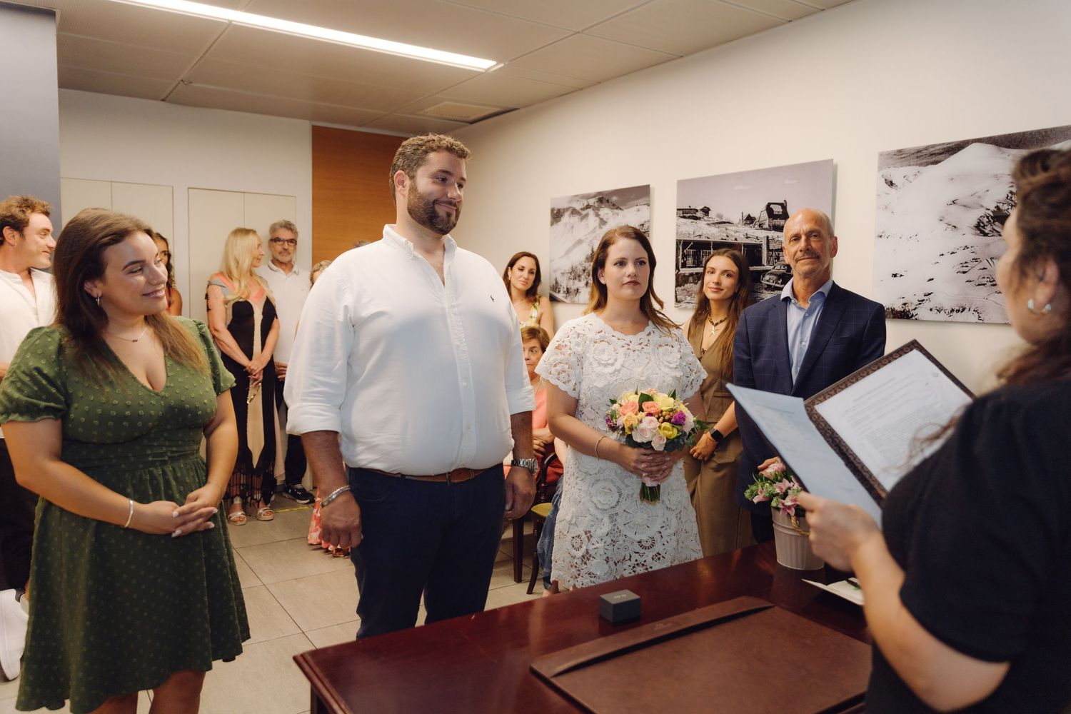 Couple stands before officiant during intimate civil wedding ceremony in small room with guests watching.