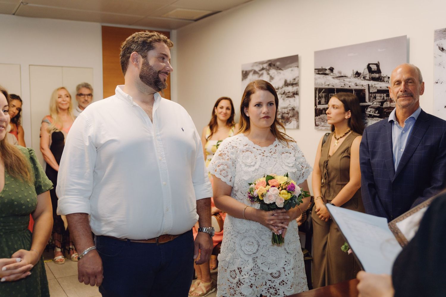 Groom in white shirt and bride in lace dress listen attentively during civil wedding ceremony with guests behind them.