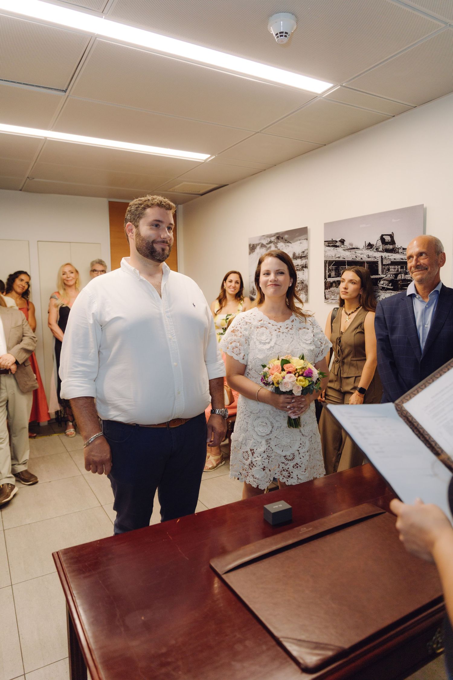 Bride and groom stand before officiant holding book during small civil wedding ceremony with family and friends gathered.
