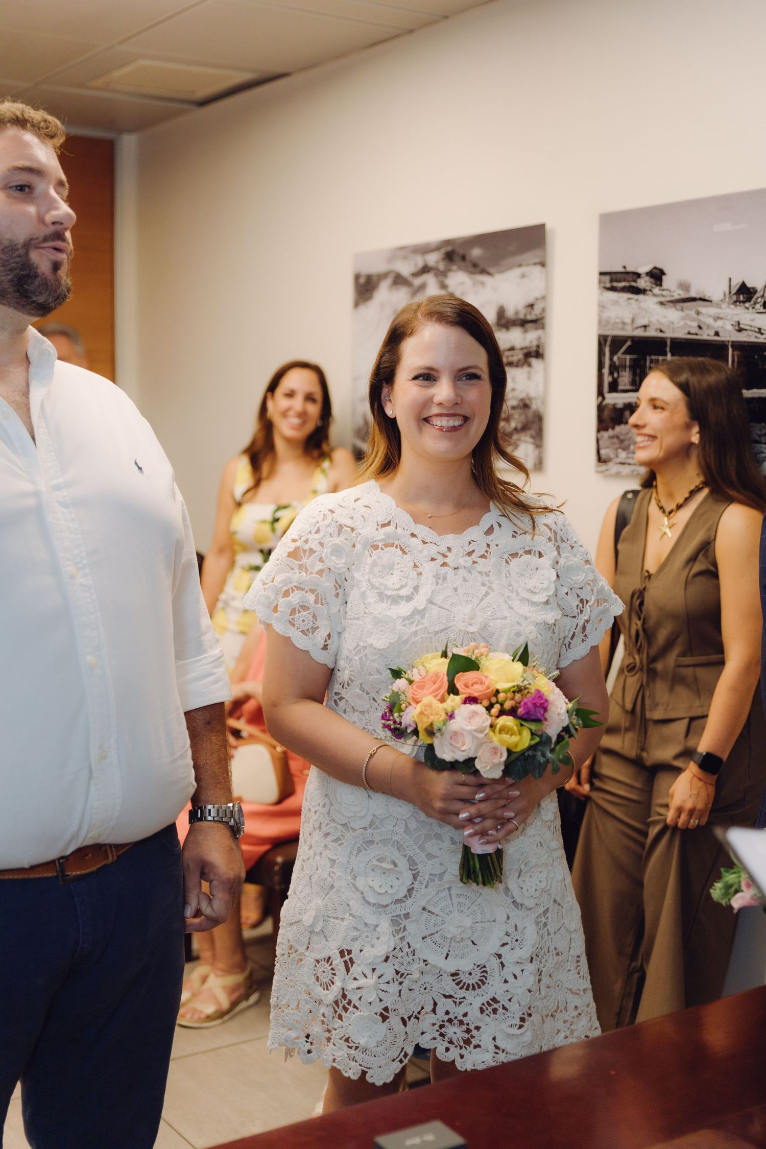 Smiling bride in white lace dress holds colorful bouquet during civil wedding ceremony with guests in background.