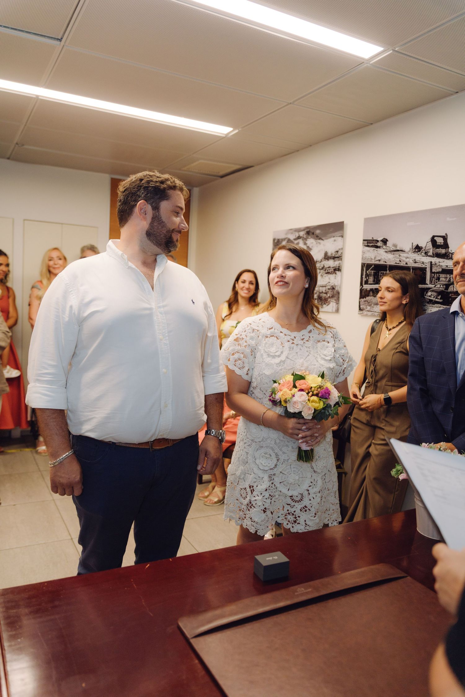Bride and groom stand together at wooden desk during intimate civil wedding ceremony surrounded by smiling guests.