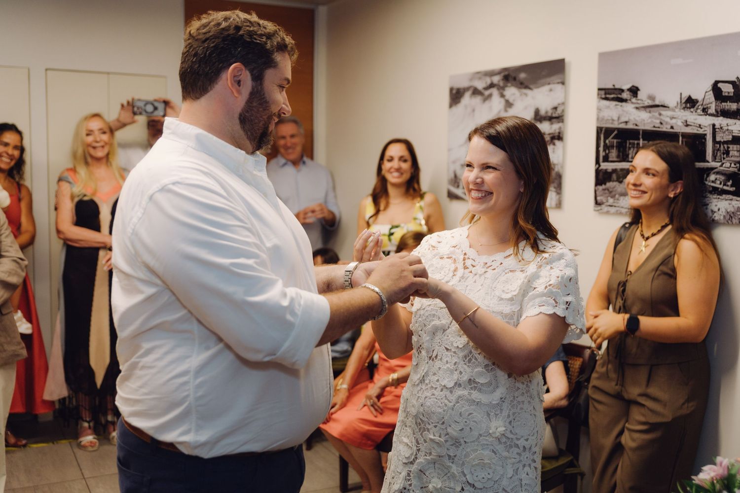 Bride and groom exchange rings and smile joyfully during civil wedding ceremony with guests celebrating behind them.