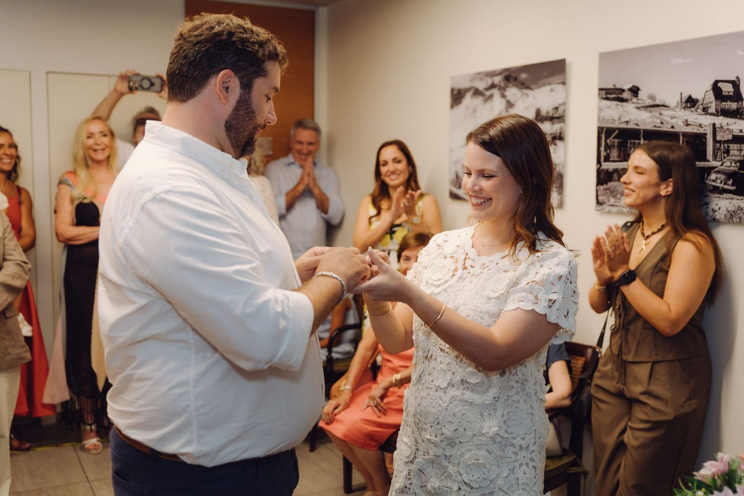 Groom places ring on brides finger as guests clap and cheer during intimate civil wedding ceremony.