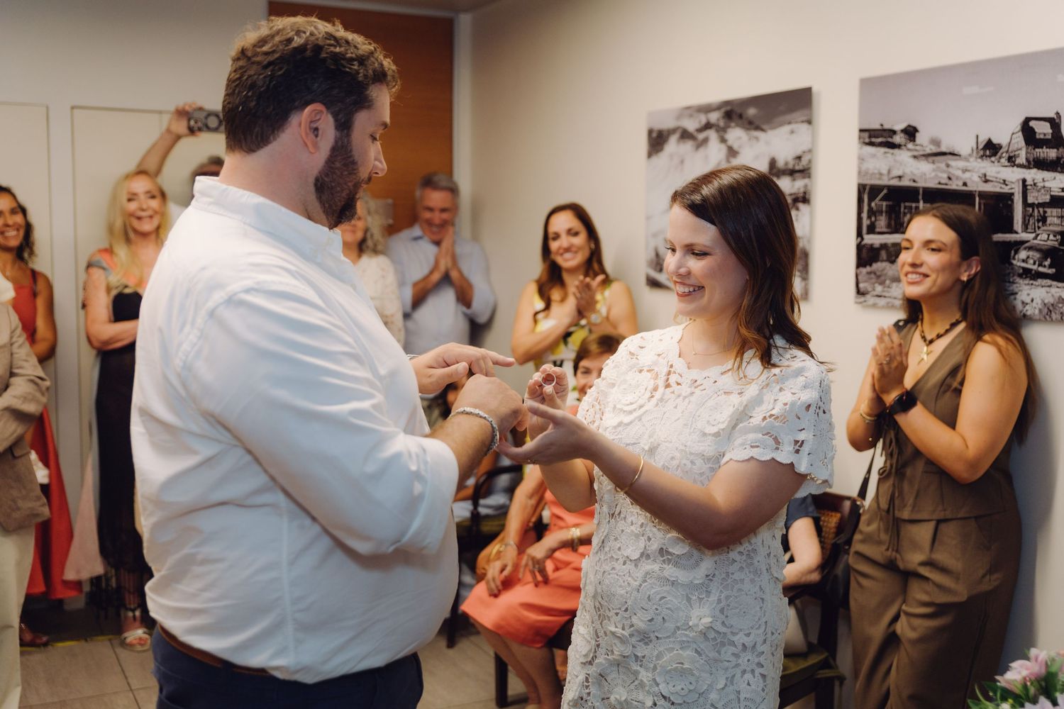 Couple exchanging rings during an intimate indoor wedding ceremony with smiling guests applauding in the background.