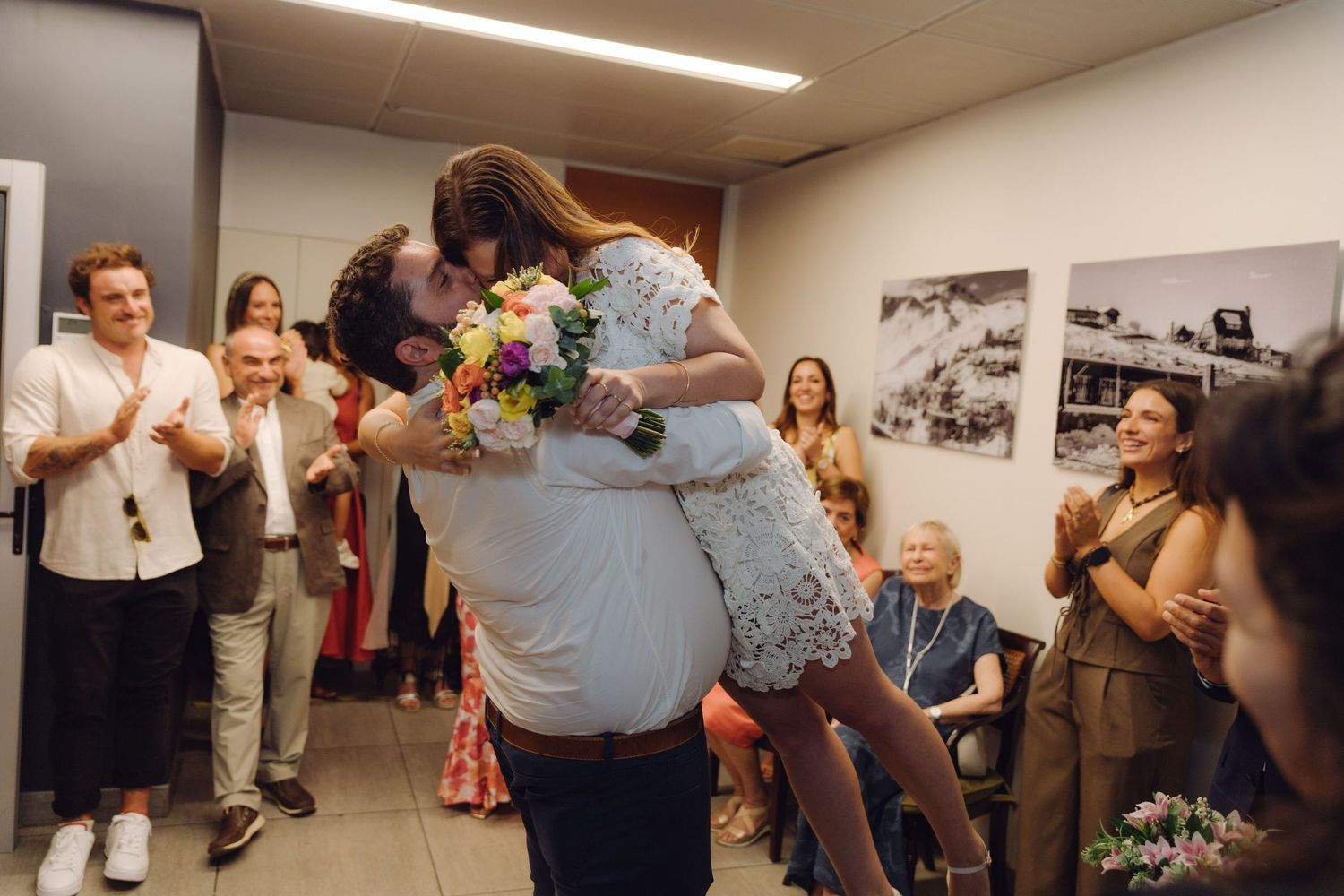 Groom lifting bride for a kiss during wedding ceremony while guests clap, bride holding colorful bouquet.