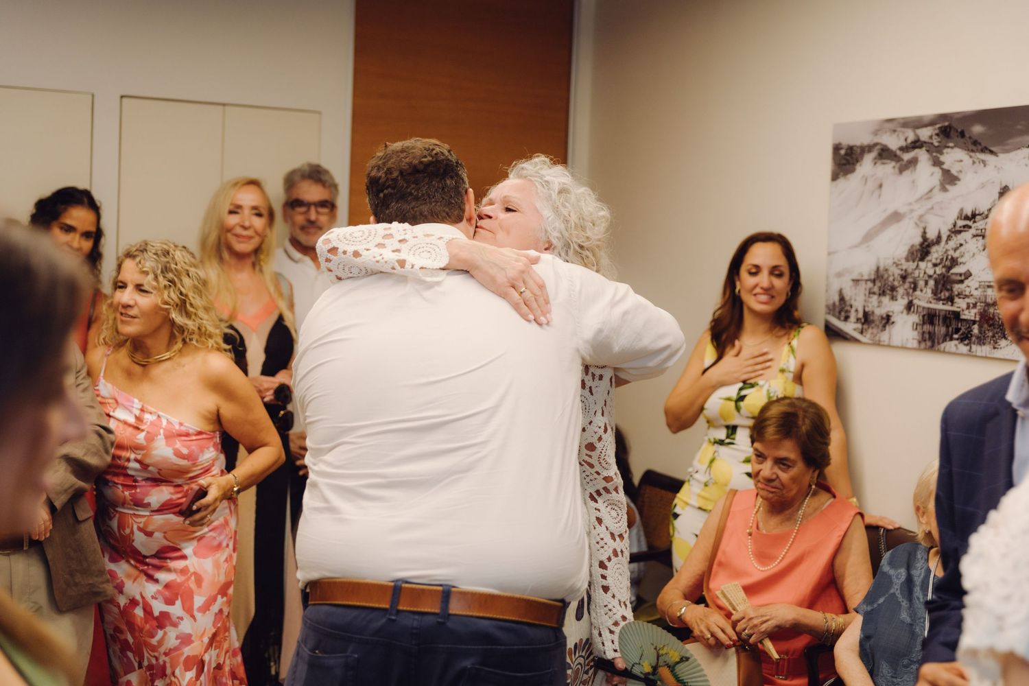 Emotional wedding moment as groom embraces older woman, likely mother, surrounded by celebrating guests indoors.
