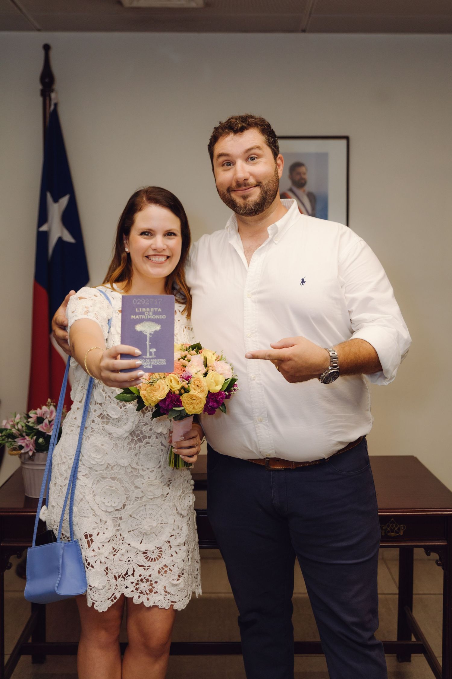 Smiling newlyweds displaying their marriage booklet and colorful bouquet at a civil wedding ceremony office.