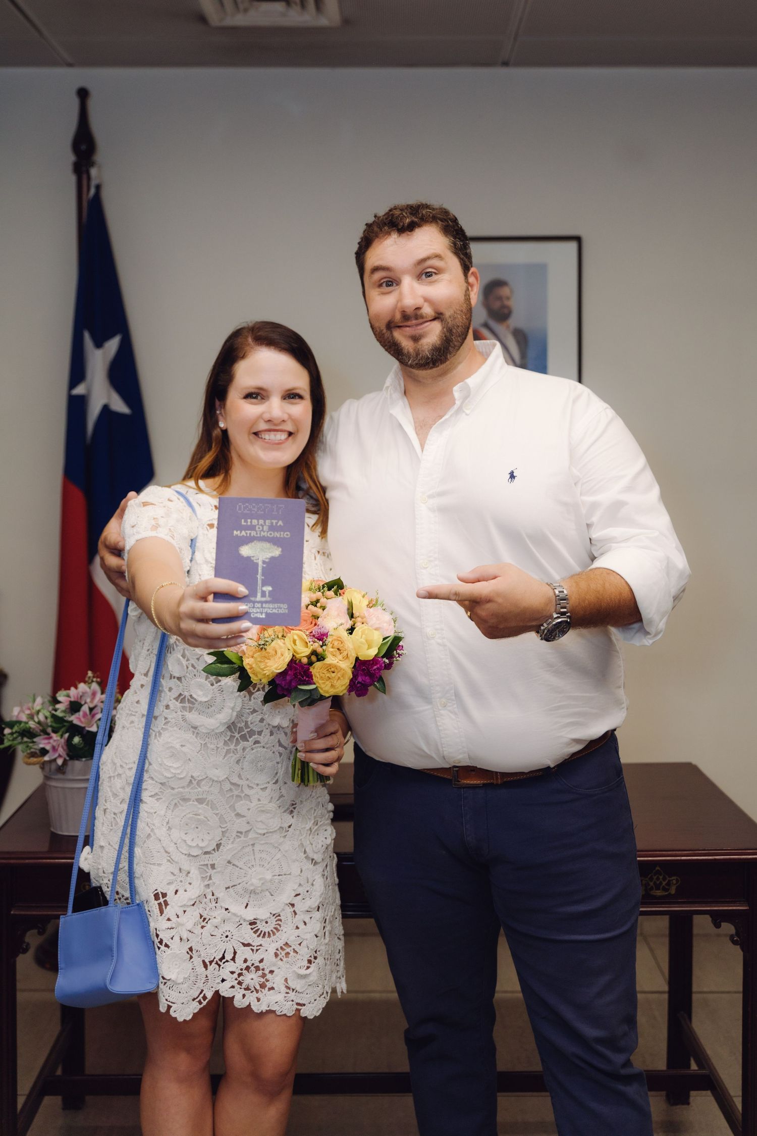 Newlywed couple posing with marriage certificate and flowers in front of Chilean flag at civil registry office.