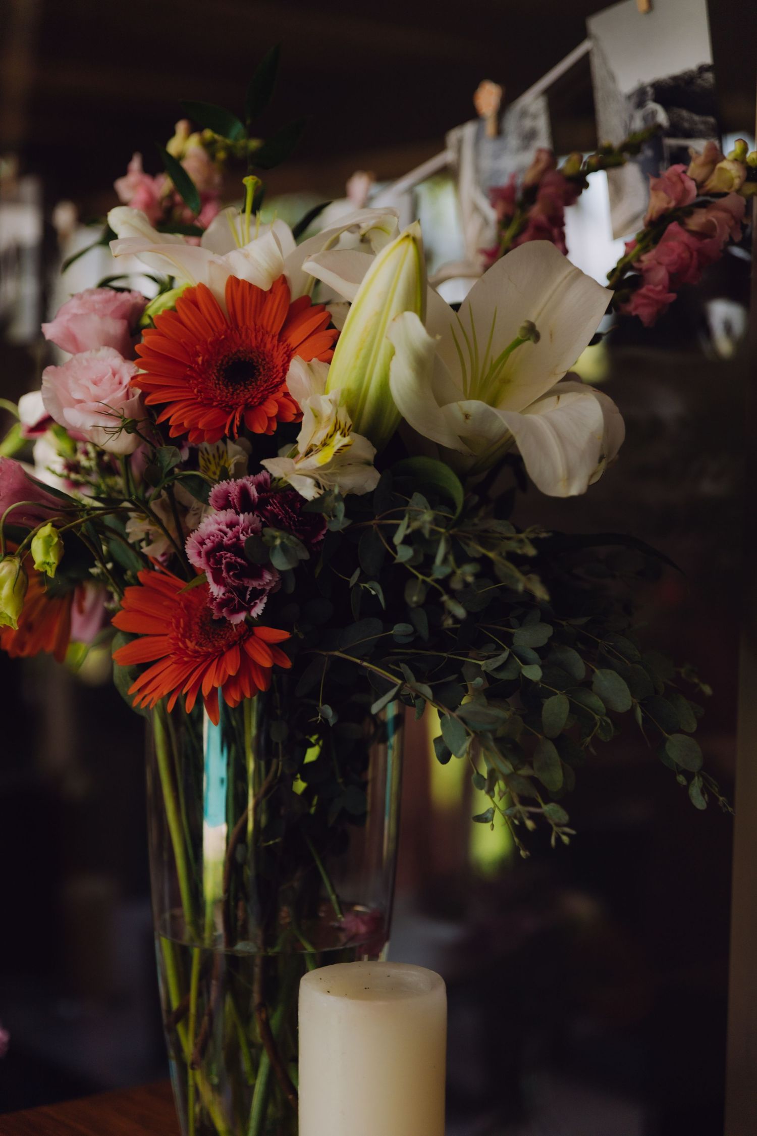 Vibrant wedding floral arrangement featuring red gerberas, white lilies, and pink roses in glass vase with candle.