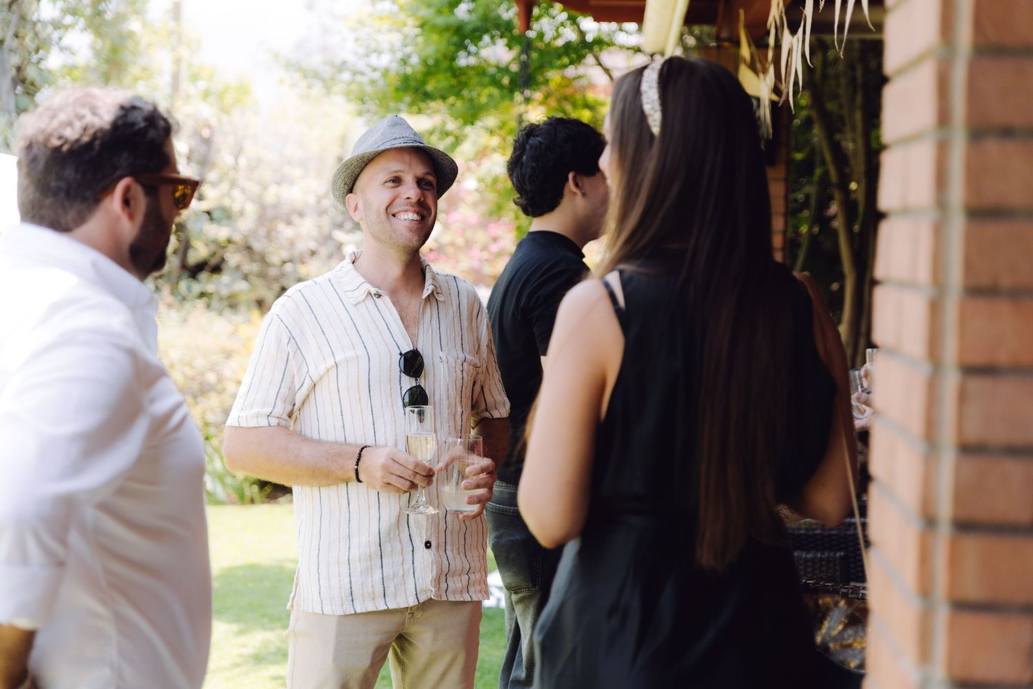 Group of guests socializing outdoors at a sunny wedding reception, man in hat holding a drink and smiling.