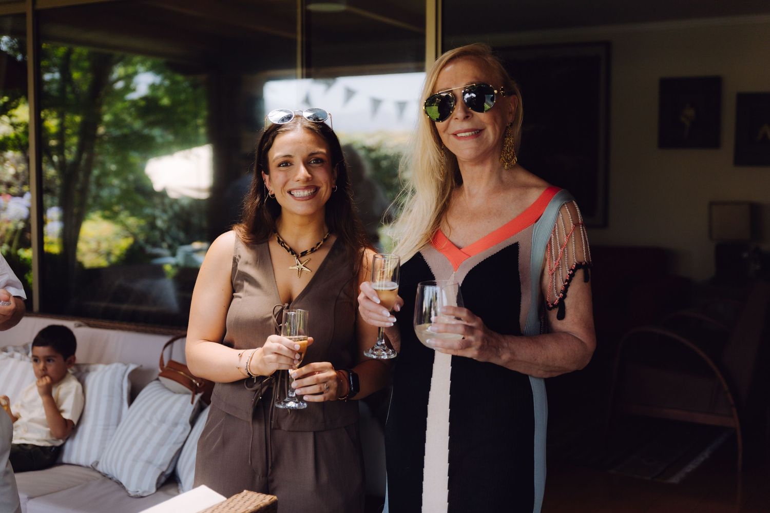 Two women smiling and holding champagne glasses at an outdoor wedding reception on a sunny day.