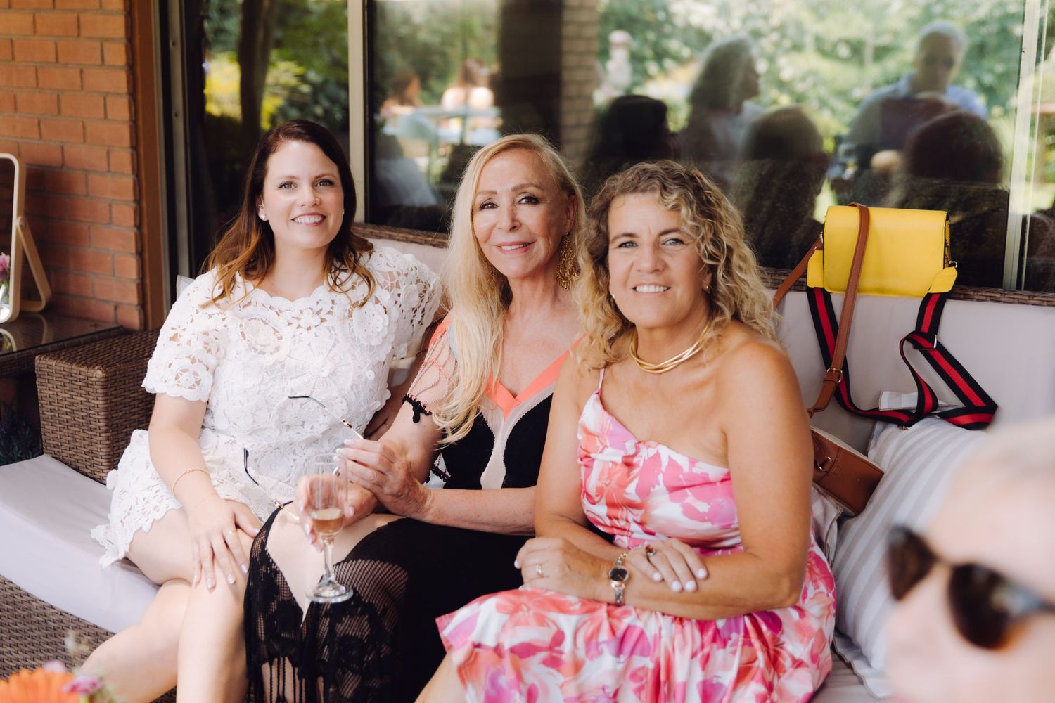Three women sitting together smiling at an outdoor wedding celebration, holding wine glasses in summer attire.