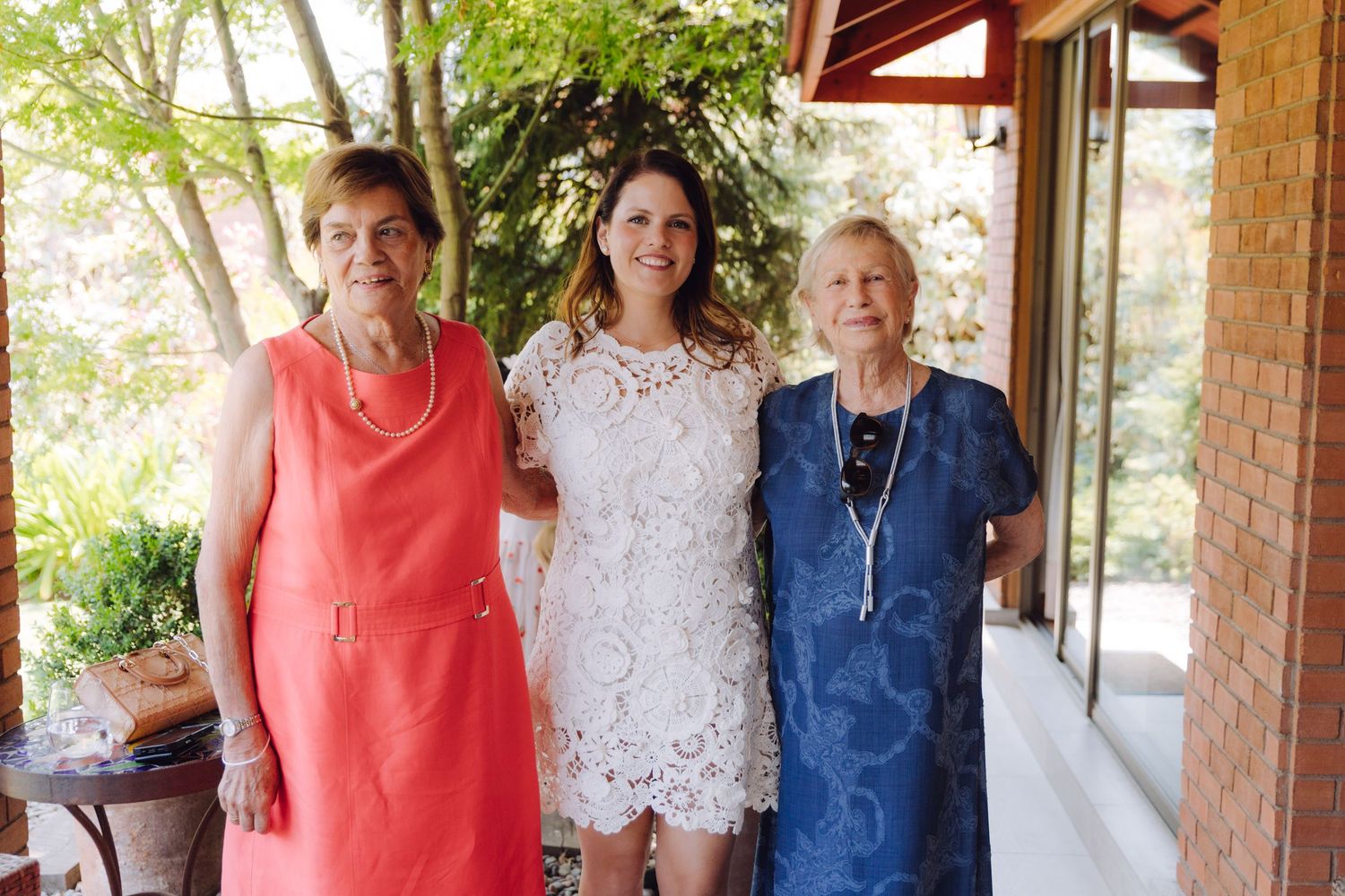 Three women in elegant dresses pose together smiling on a sunlit porch.
