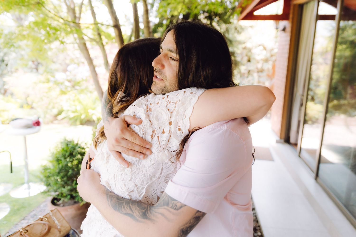 Two women in a warm embrace outdoors near a sunlit porch.