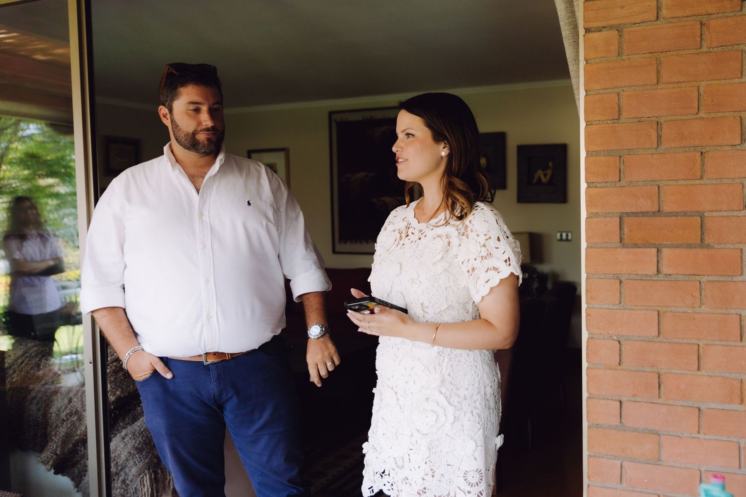 A man in white shirt and woman in lace dress stand talking in a doorway.