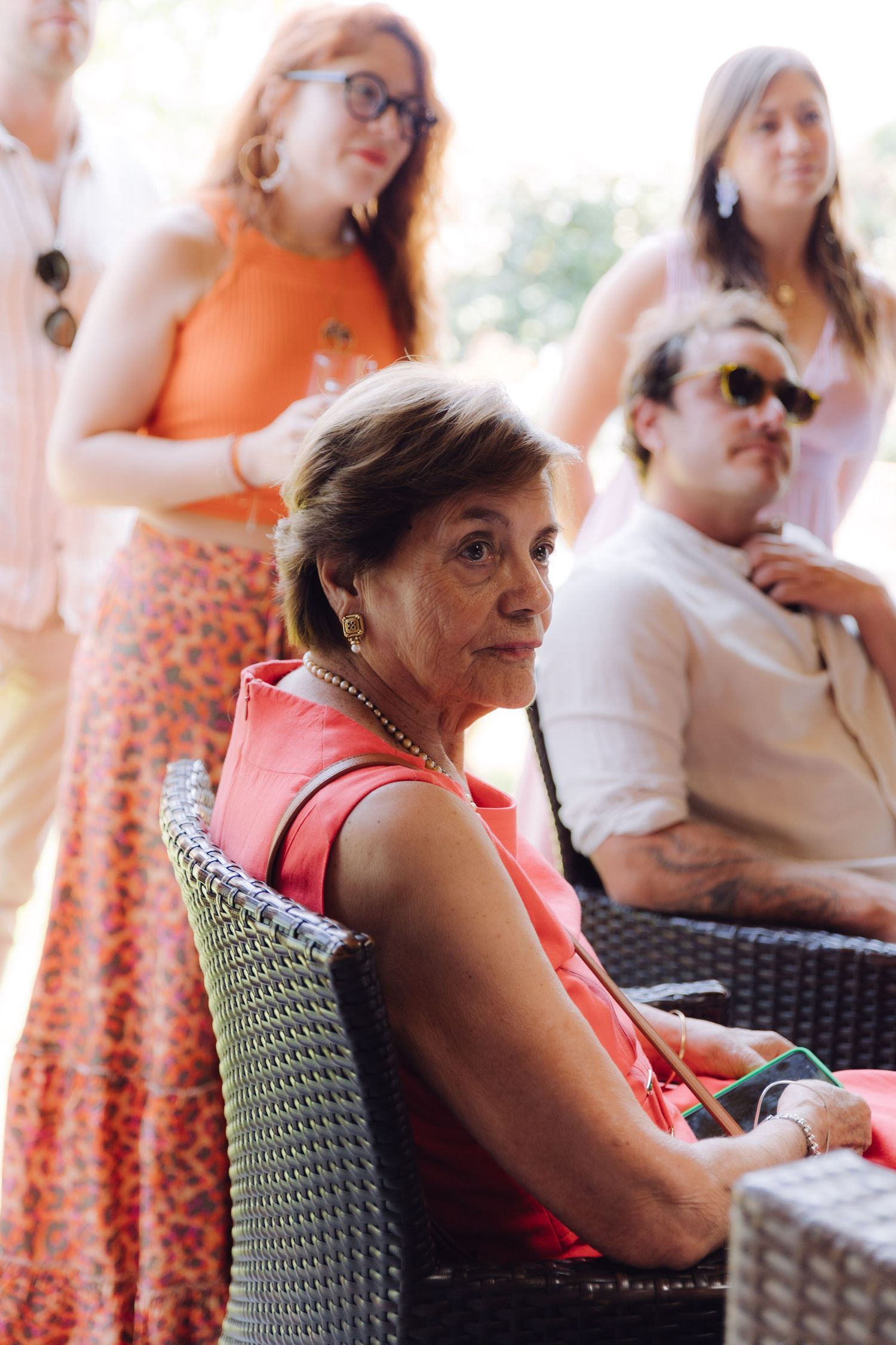 An older woman in coral dress sits attentively at an outdoor gathering.