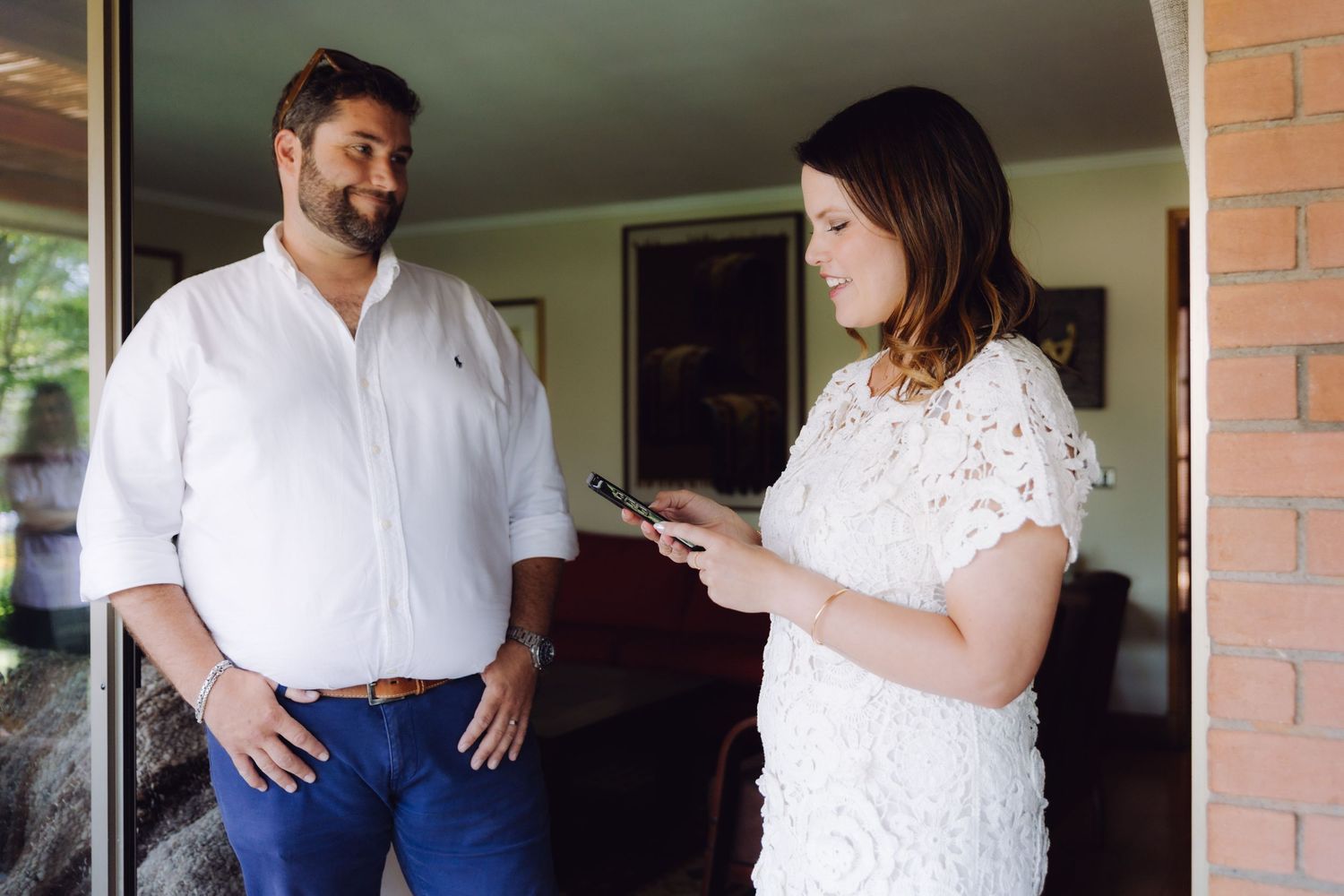A man in white shirt watches a woman in lace dress reading from a phone.