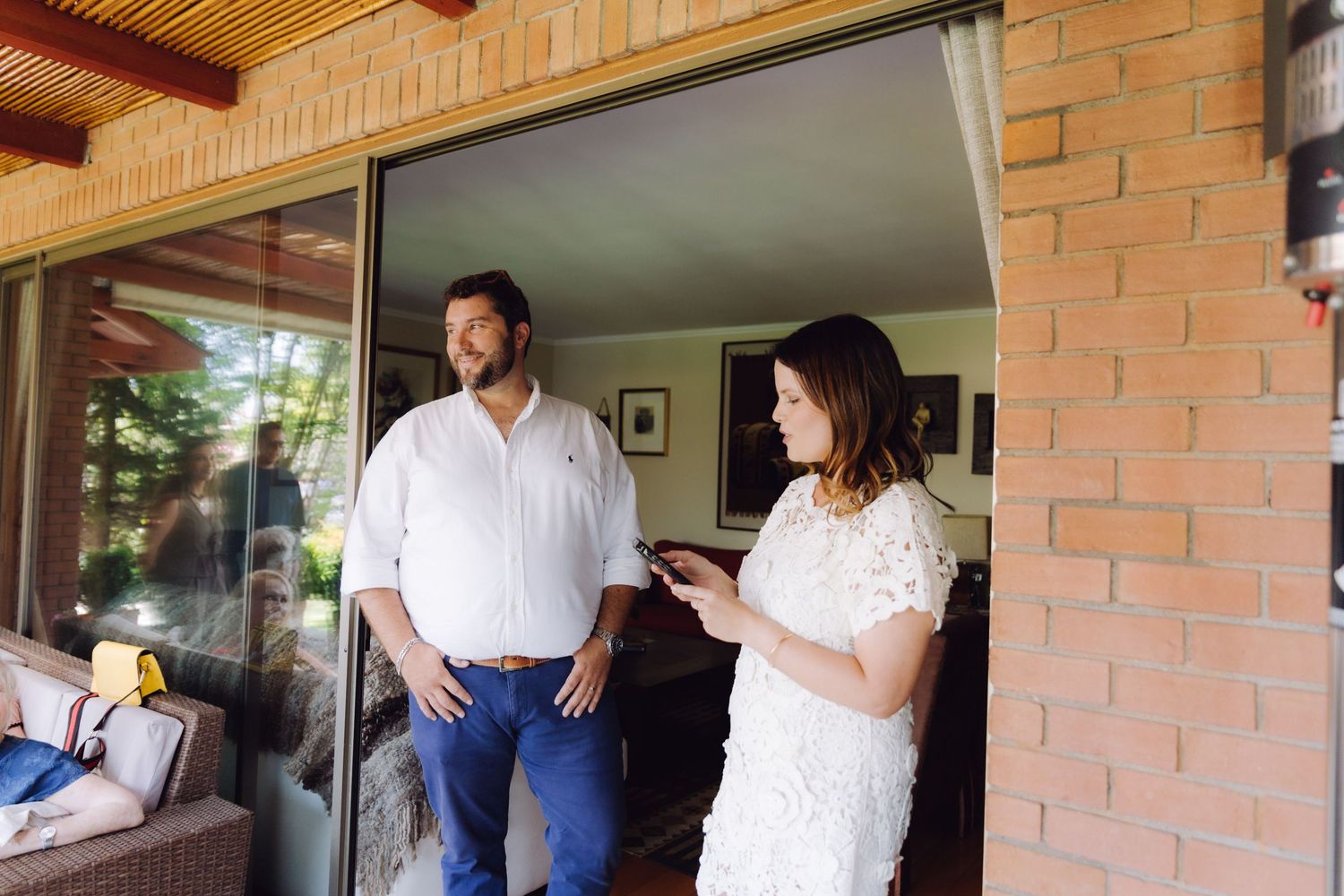 A couple stands on a brick porch, woman reading from phone, man watching.