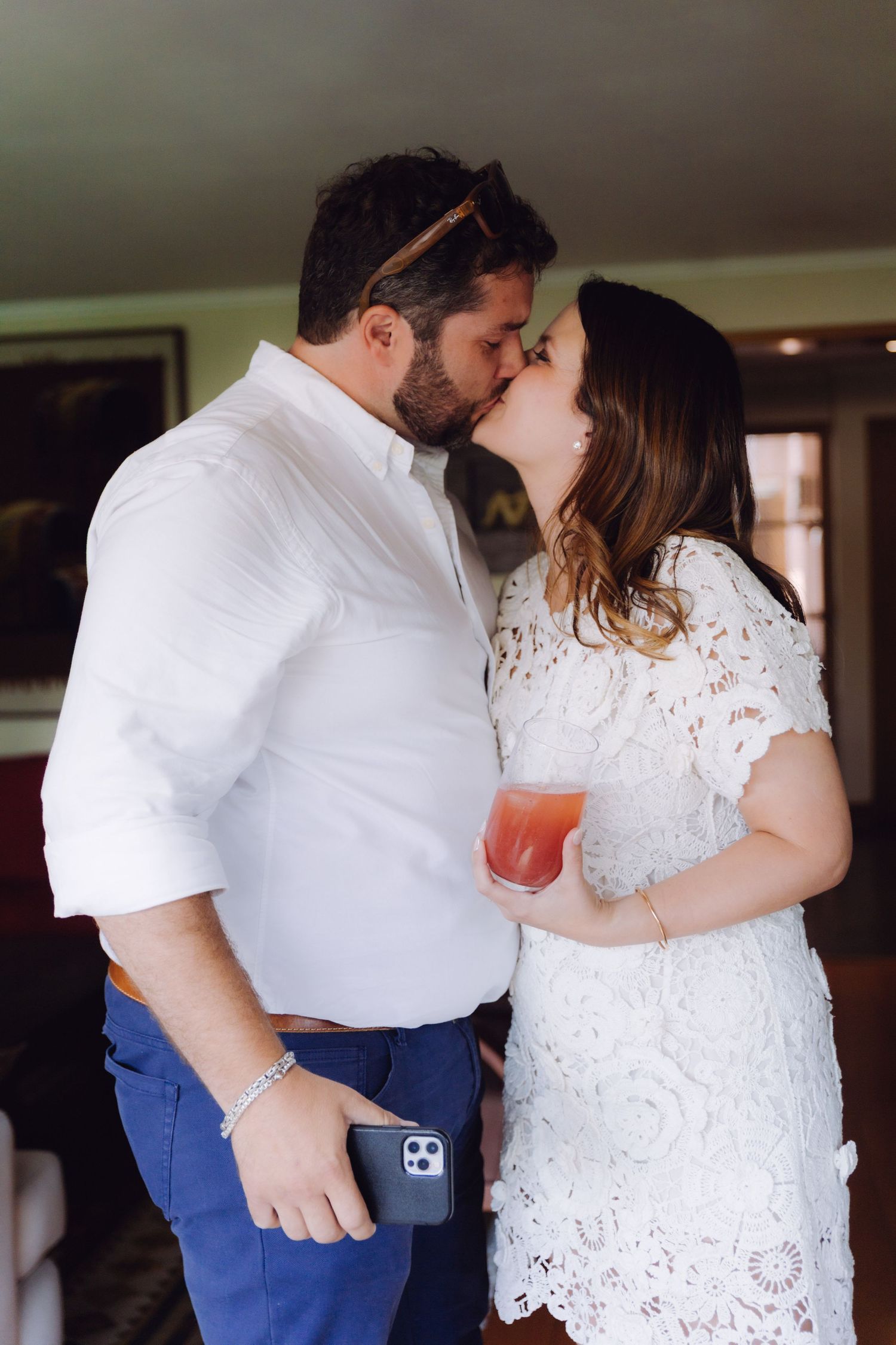 Couple sharing a romantic kiss at a wedding celebration, woman in white lace dress holding a red cocktail.