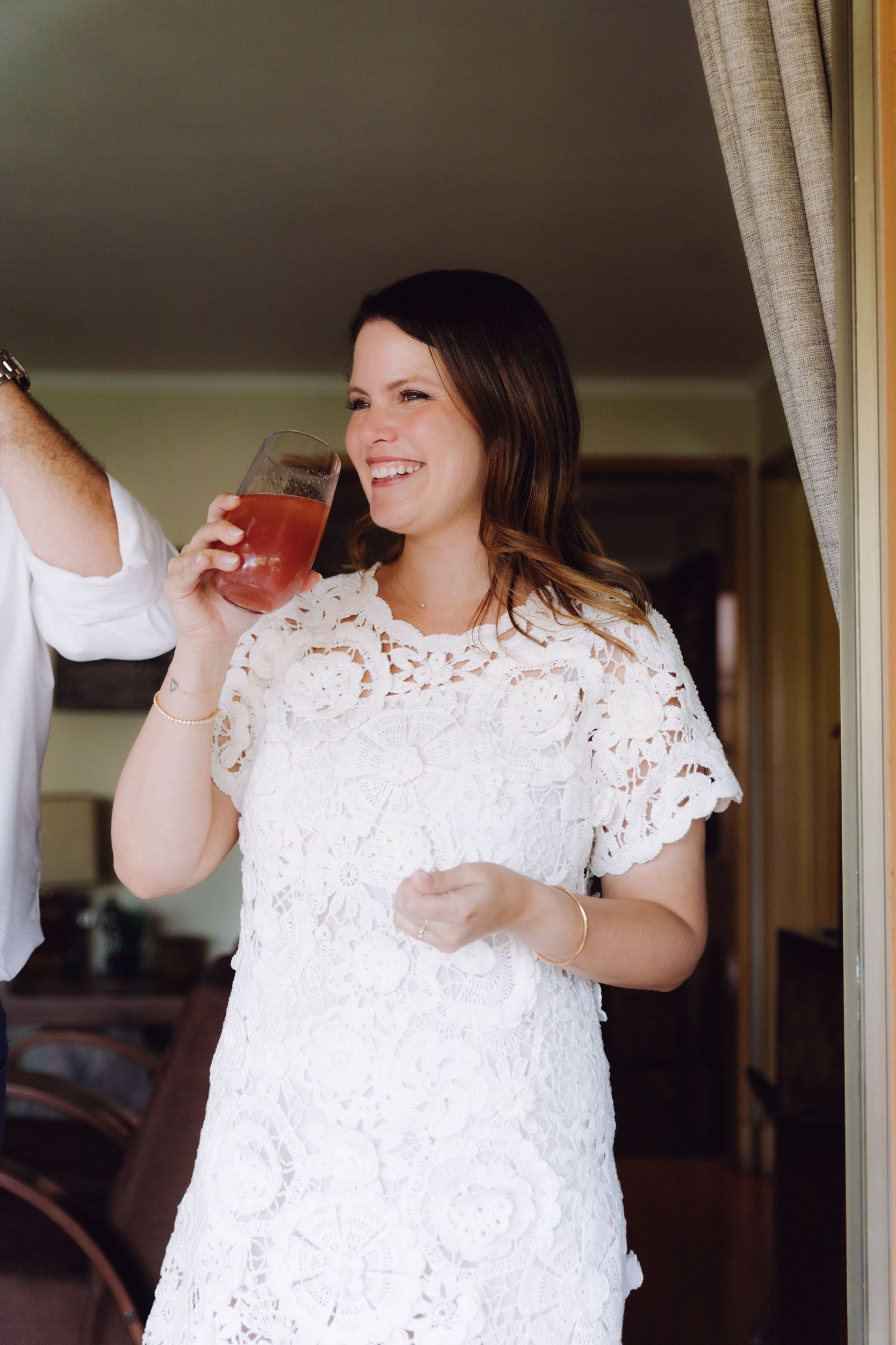 Smiling bride in white lace dress sipping a red cocktail at her wedding reception.