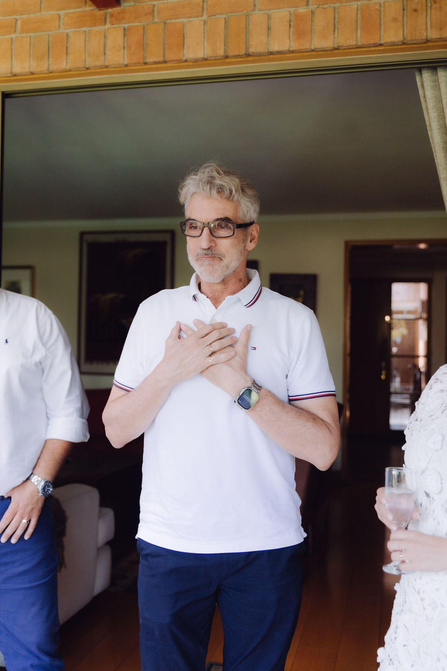Older man in white polo shirt with glasses placing hands on chest in emotional gesture at wedding.