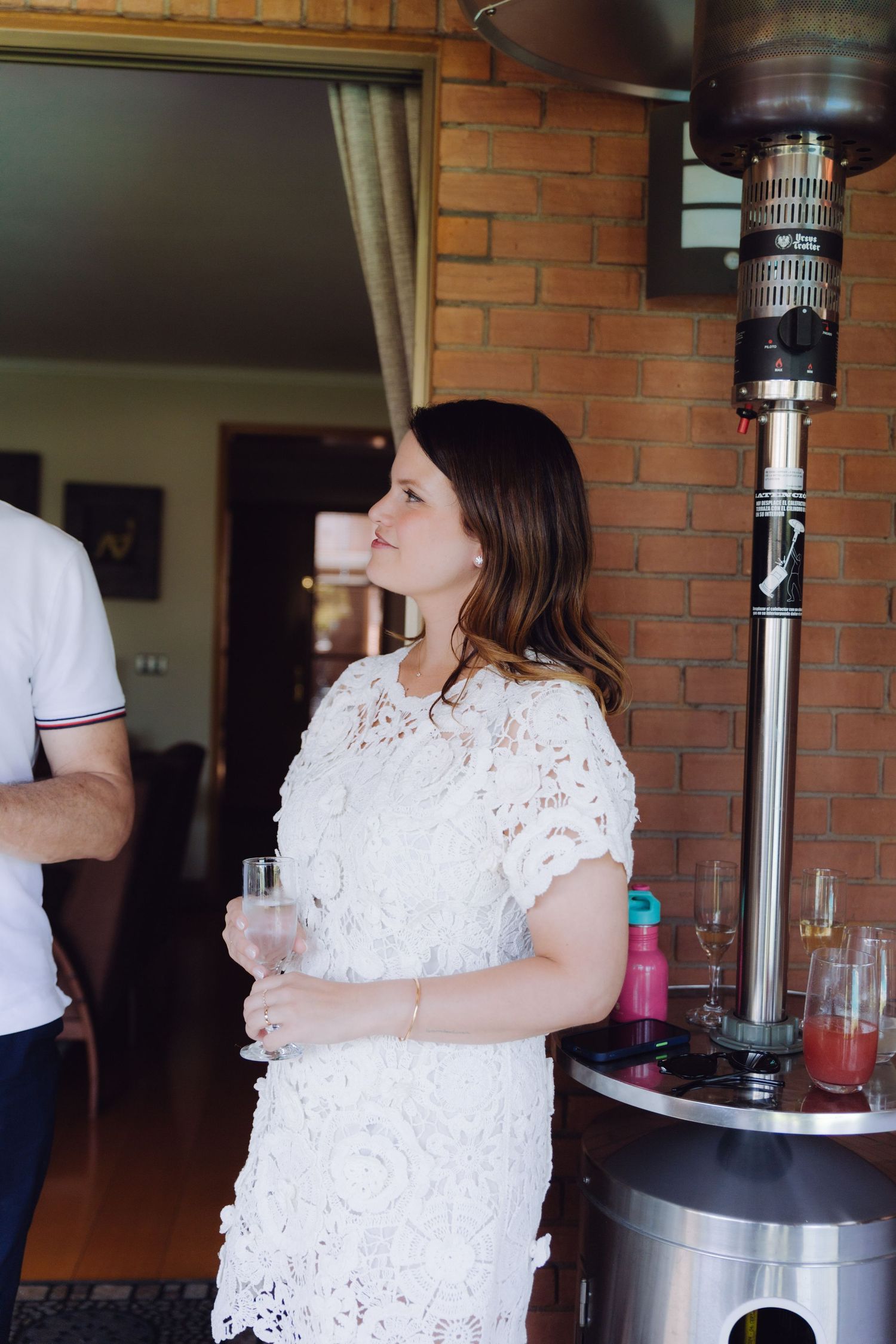 Brunette woman in white lace dress holding champagne glass at an outdoor wedding reception.