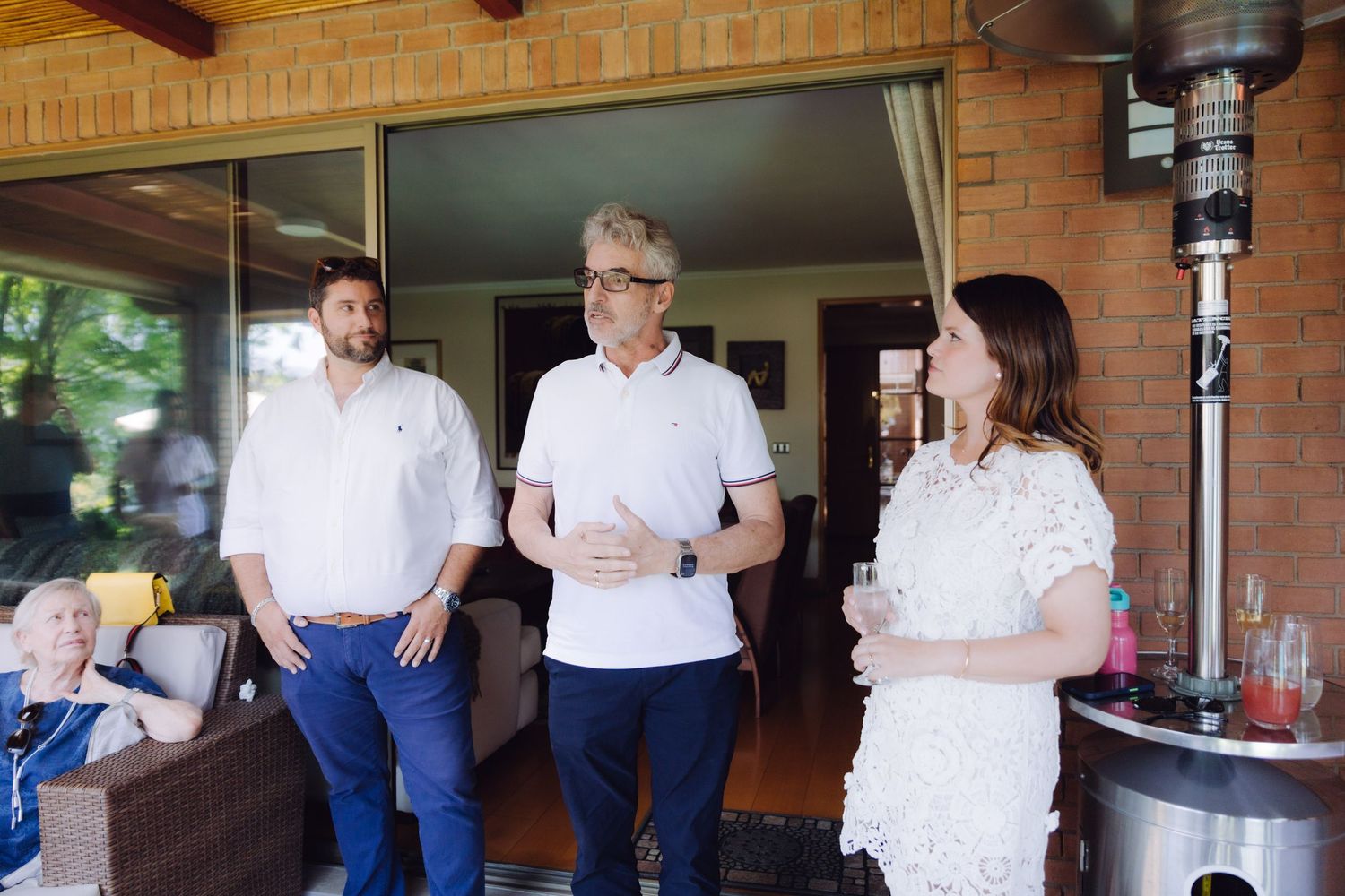 Three wedding guests conversing on a patio, two men and a woman in white lace dress.