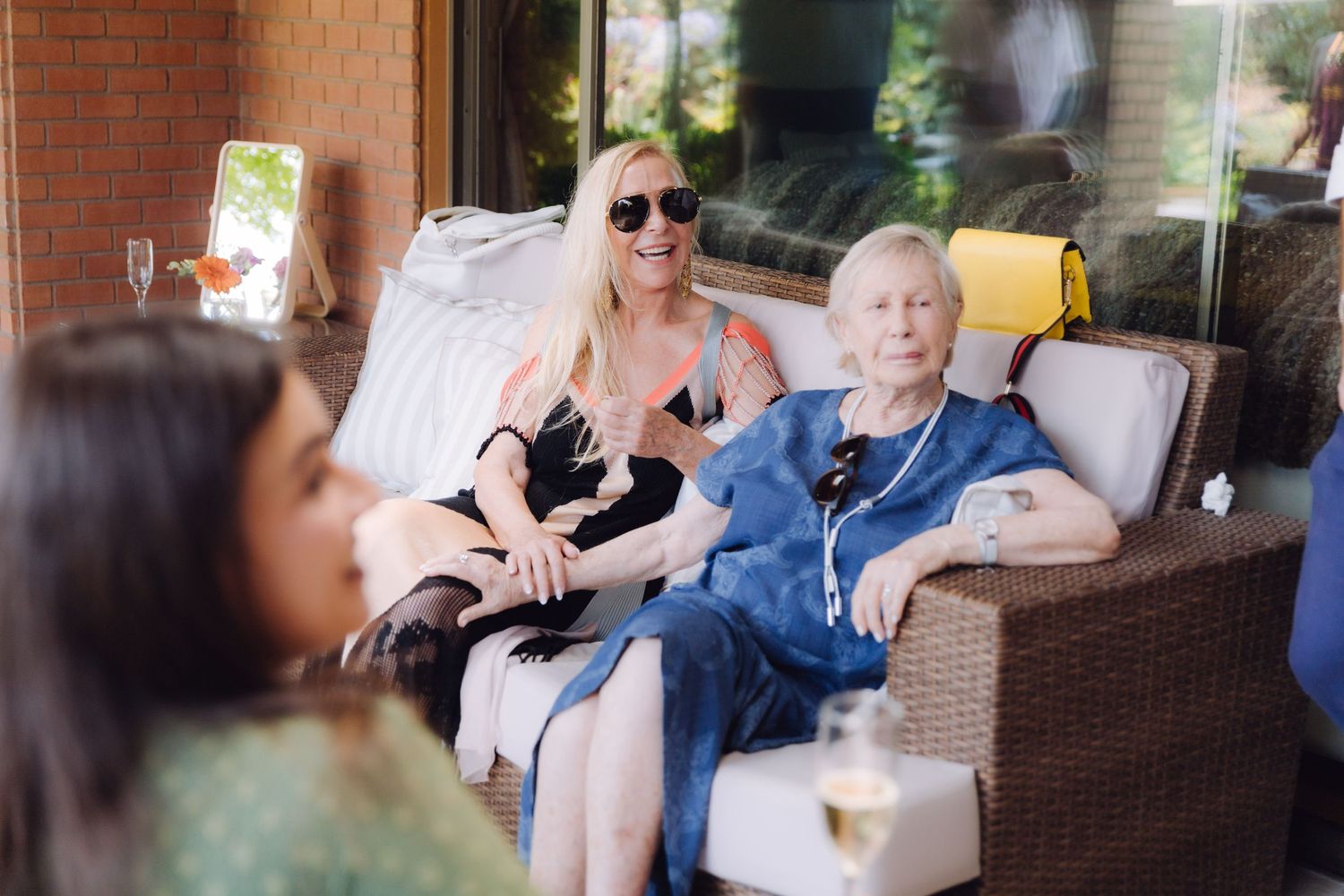 Two women seated on wicker outdoor sofa at a wedding, one wearing sunglasses holding a small dog.