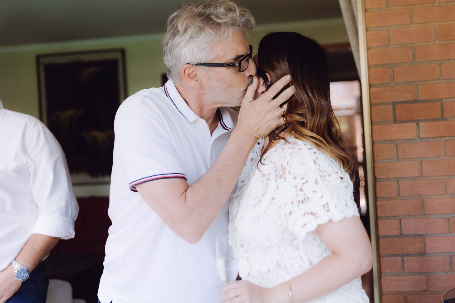 Older man in white polo gently kissing woman in white lace dress on the cheek at a wedding.