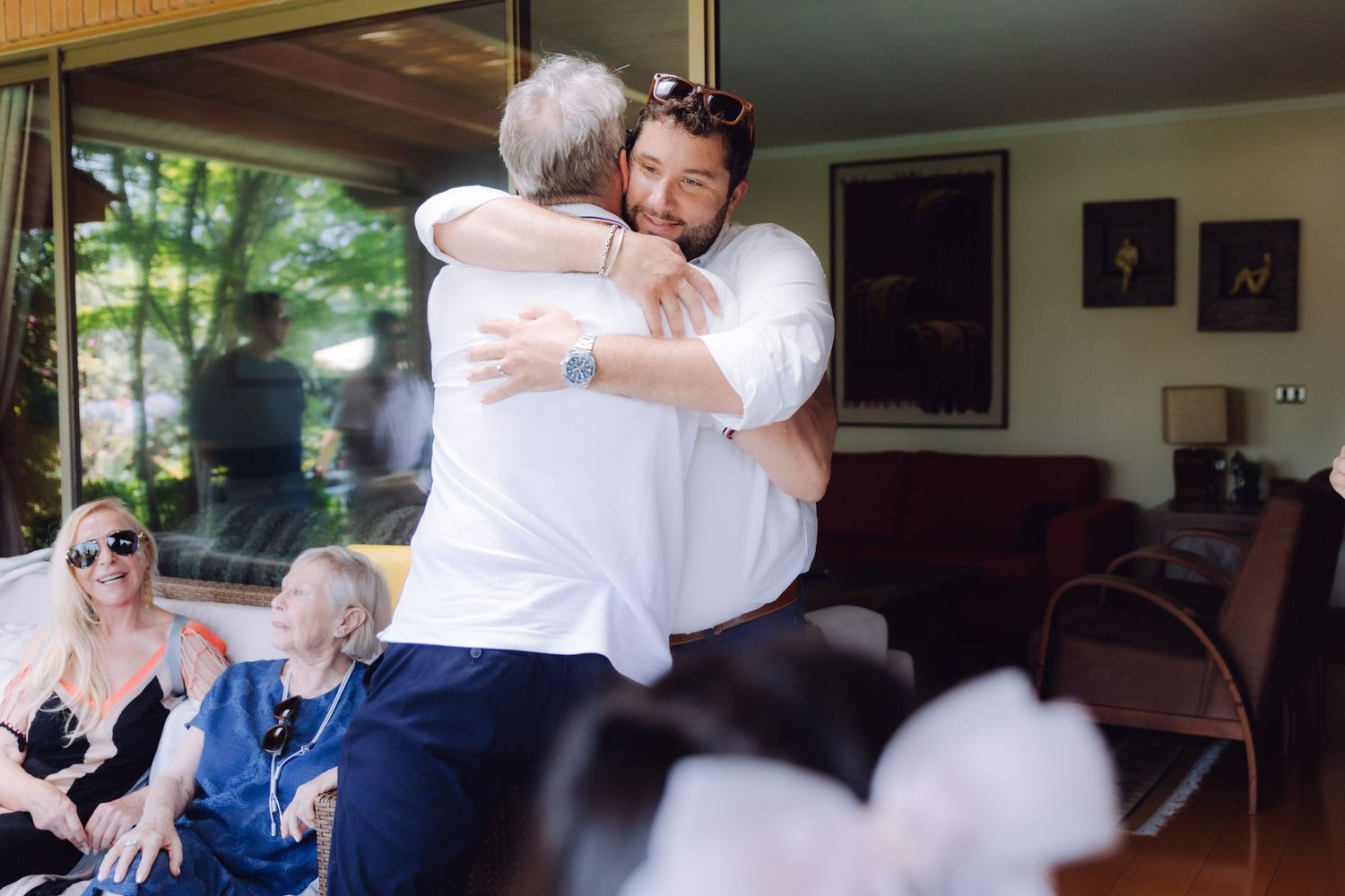 Two men sharing a warm embrace at a wedding reception with seated guests in the background.