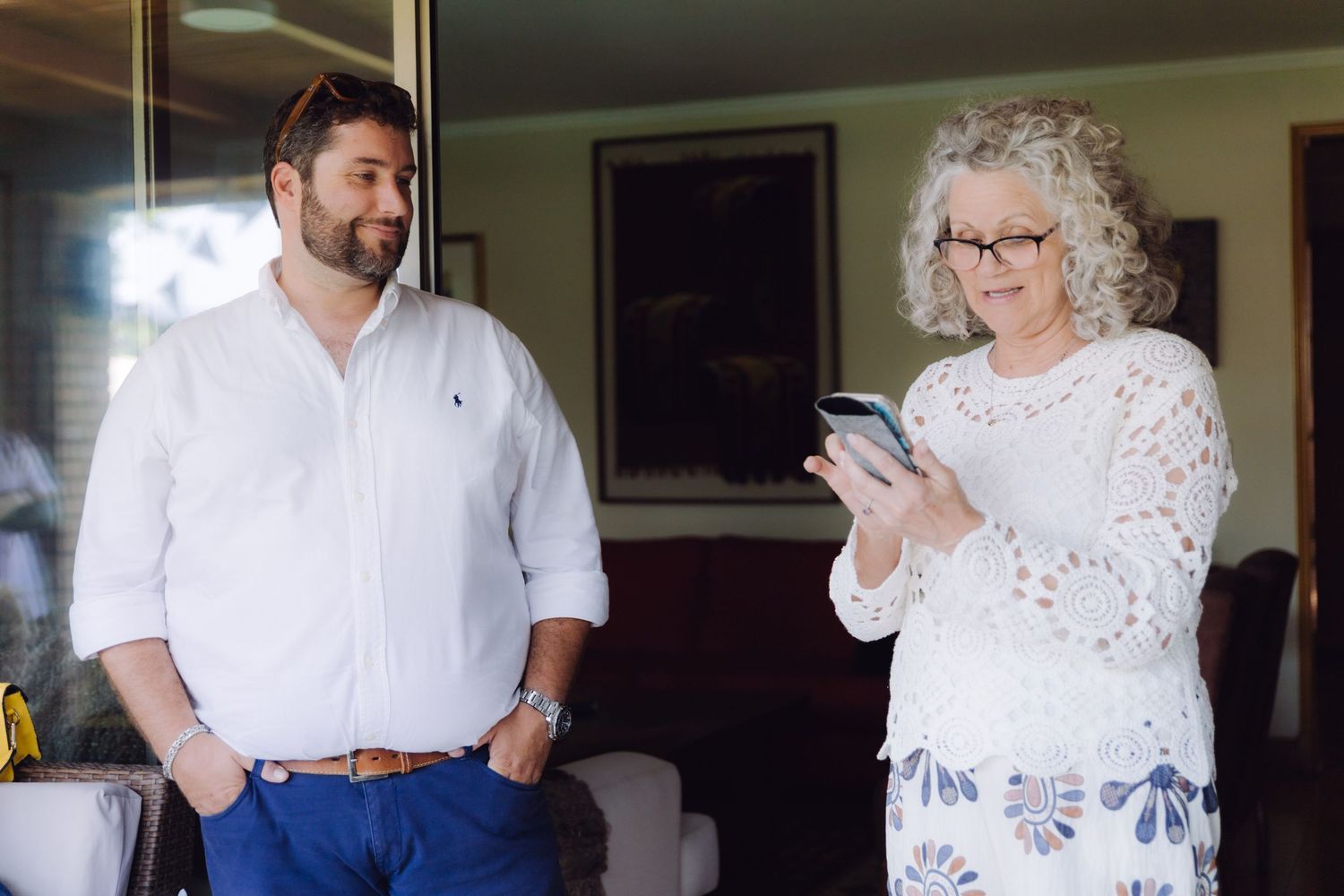 Man in white shirt standing beside older woman in lace top looking at a smartphone indoors.