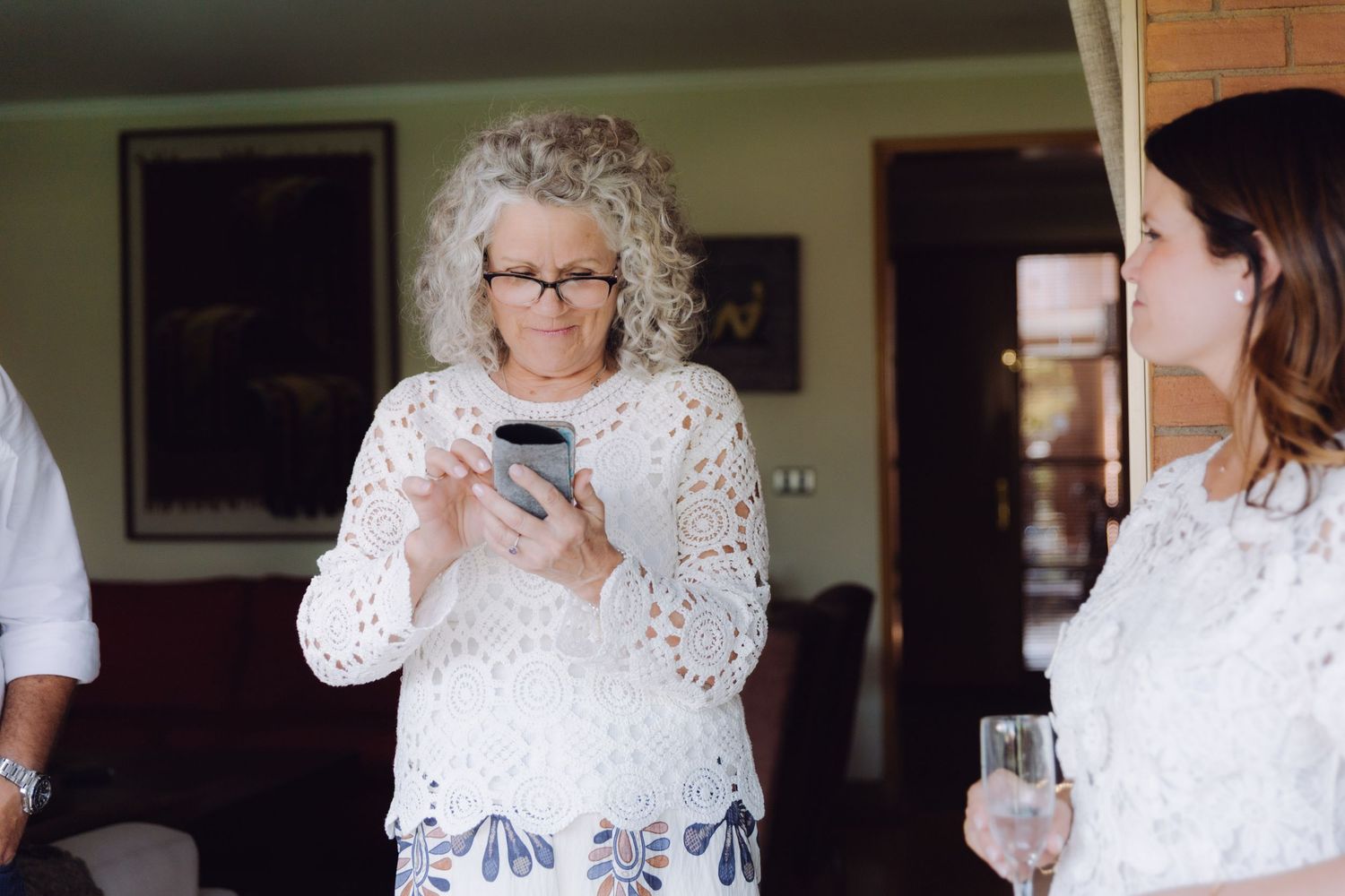Older woman with curly gray hair and glasses holding a phone at an indoor gathering.