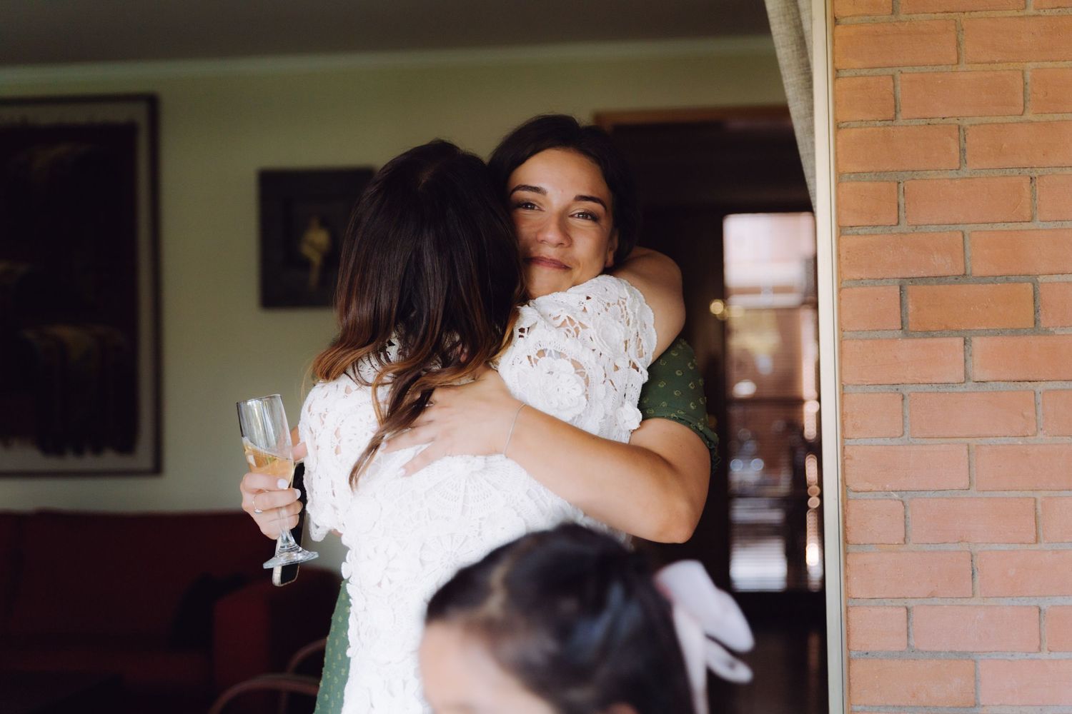 Two women sharing a warm embrace near a brick wall doorway at a wedding.