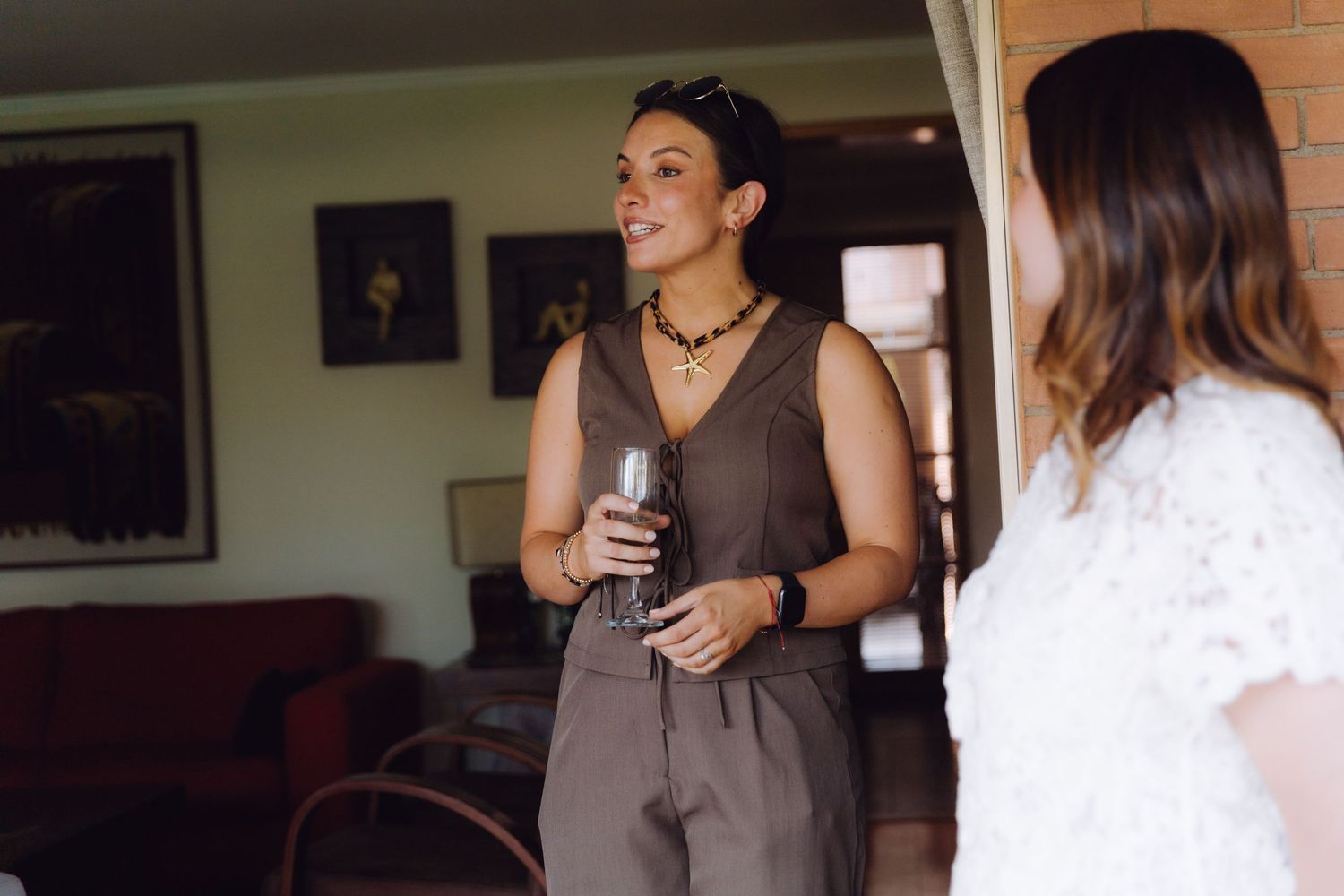 Woman in brown sleeveless outfit holding a wine glass chatting at an indoor event.