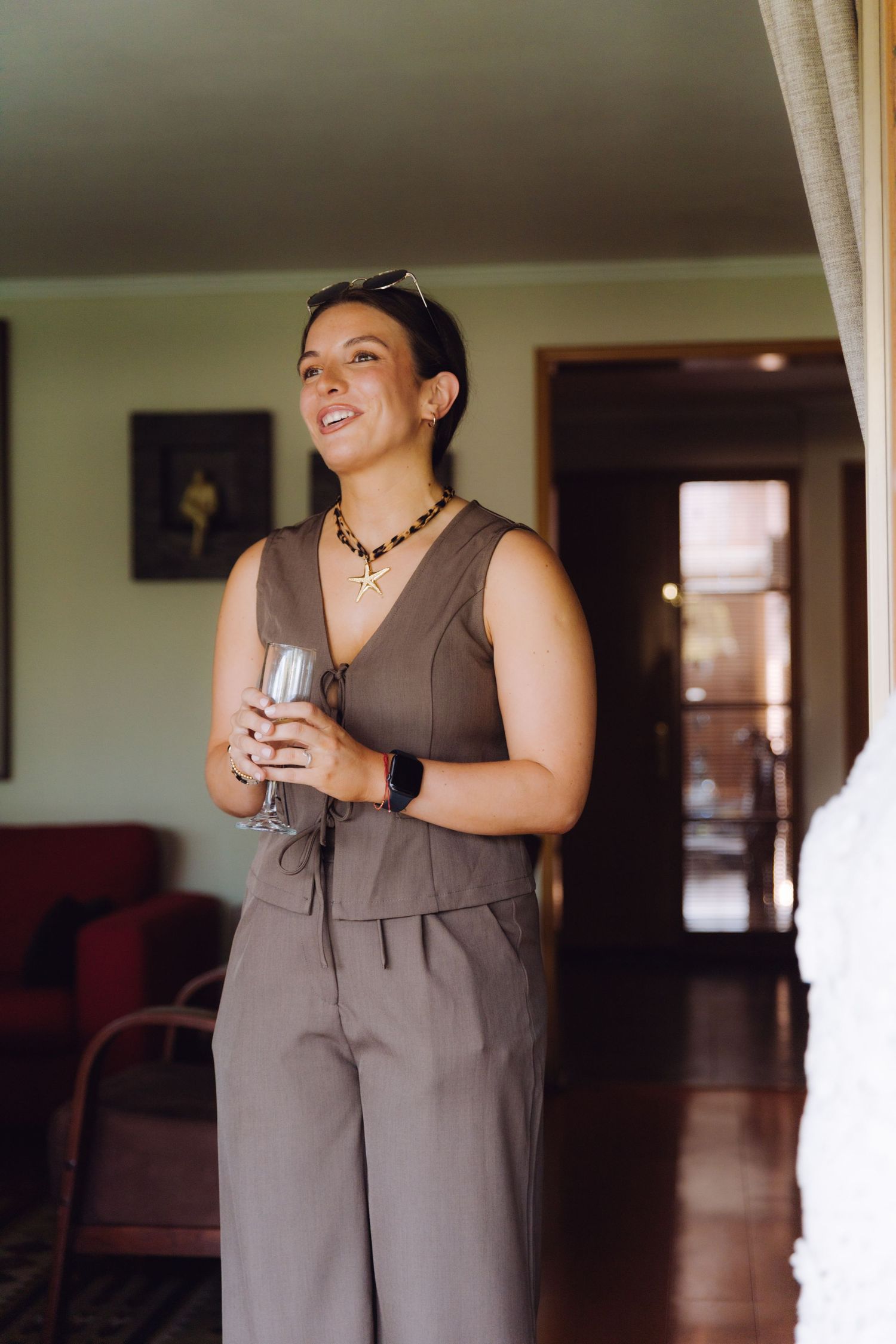 Smiling woman in brown jumpsuit holding champagne glass standing in elegant home interior.