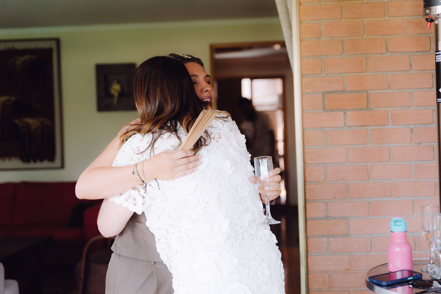 Two women hugging warmly near a brick wall while one holds a champagne glass.