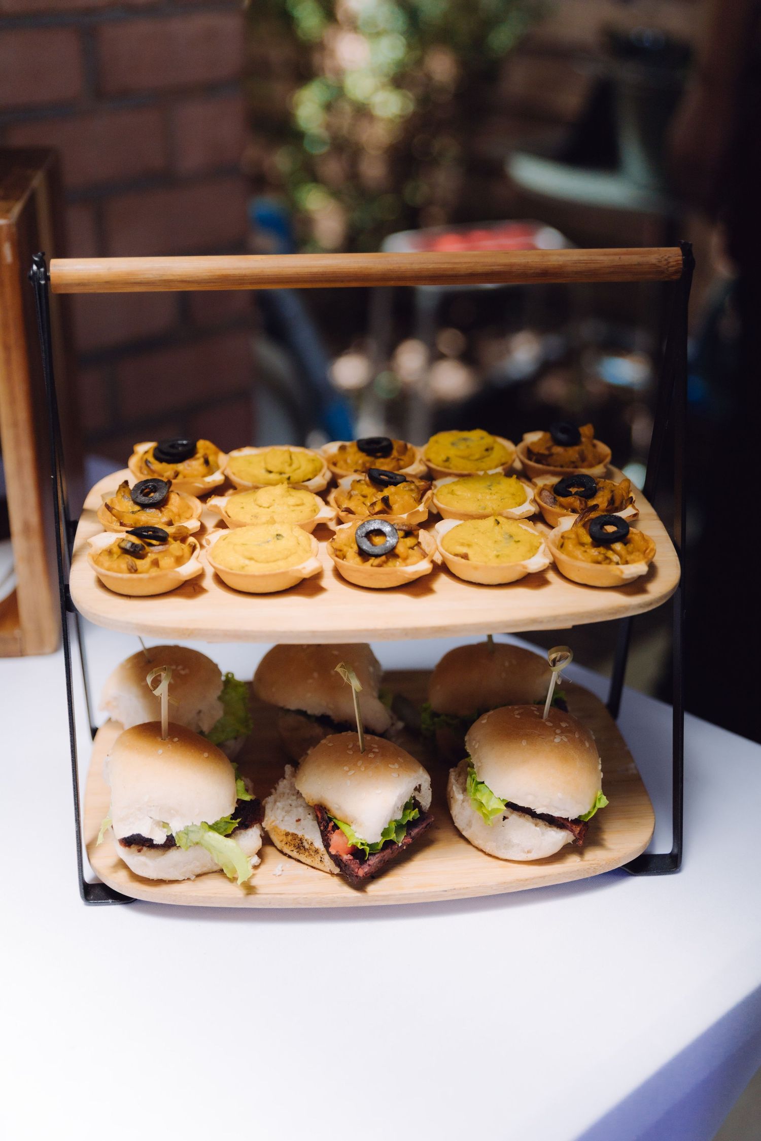 Two-tiered serving stand with mini burgers and savory canapes topped with olives at a catered event.