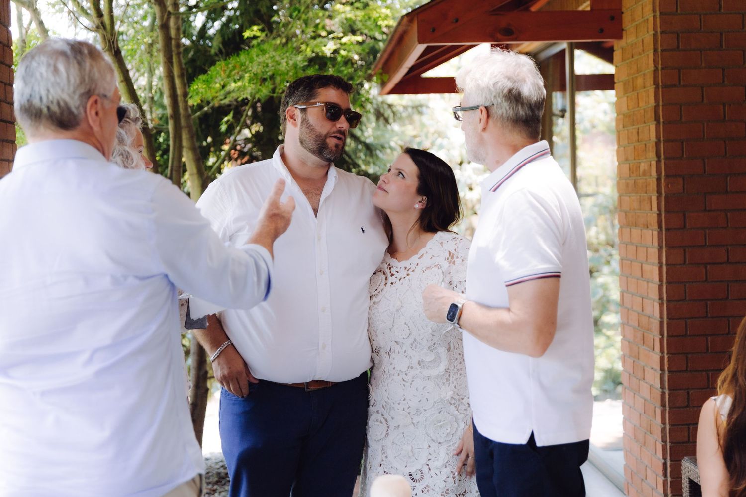 Guests socializing at an outdoor wedding reception under a shaded pergola on a sunny day.