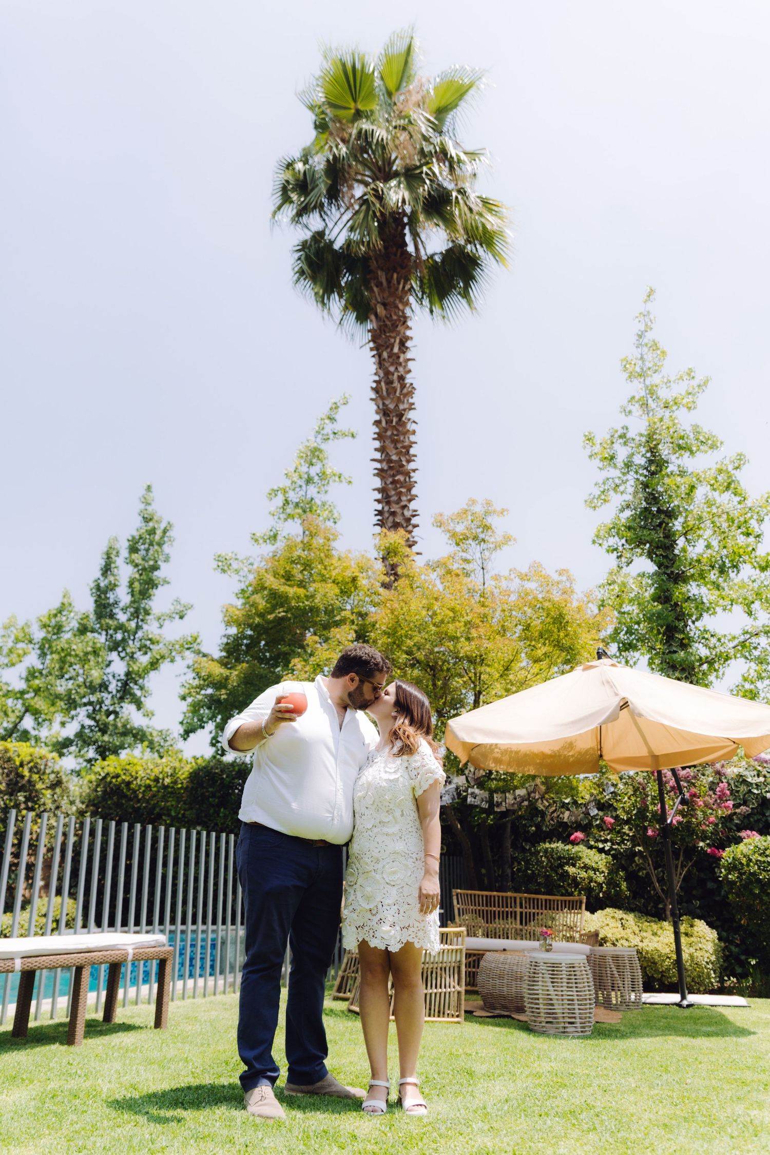 Newlyweds sharing a kiss in a garden with a tall palm tree and pool in the background.