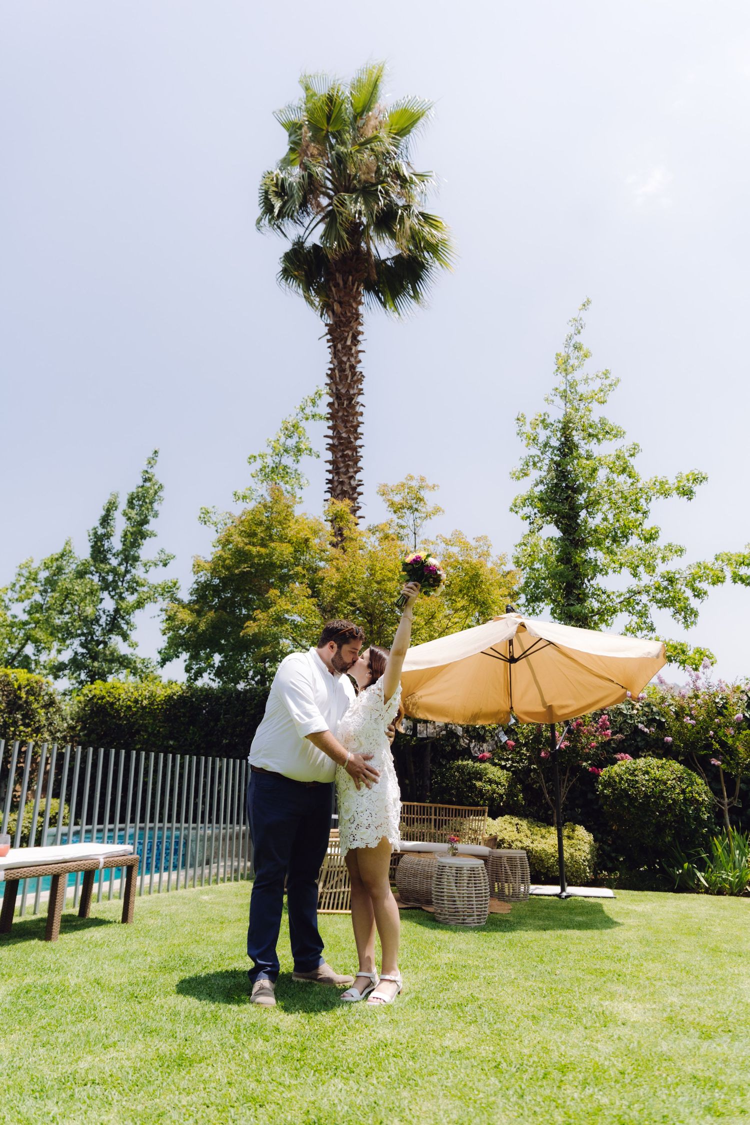 Bride and groom kissing outdoors, bride raising bouquet joyfully near a pool and palm tree.