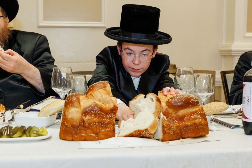Young boy in black hat and coat cutting challah bread at a Jewish Shabbat dinner table.